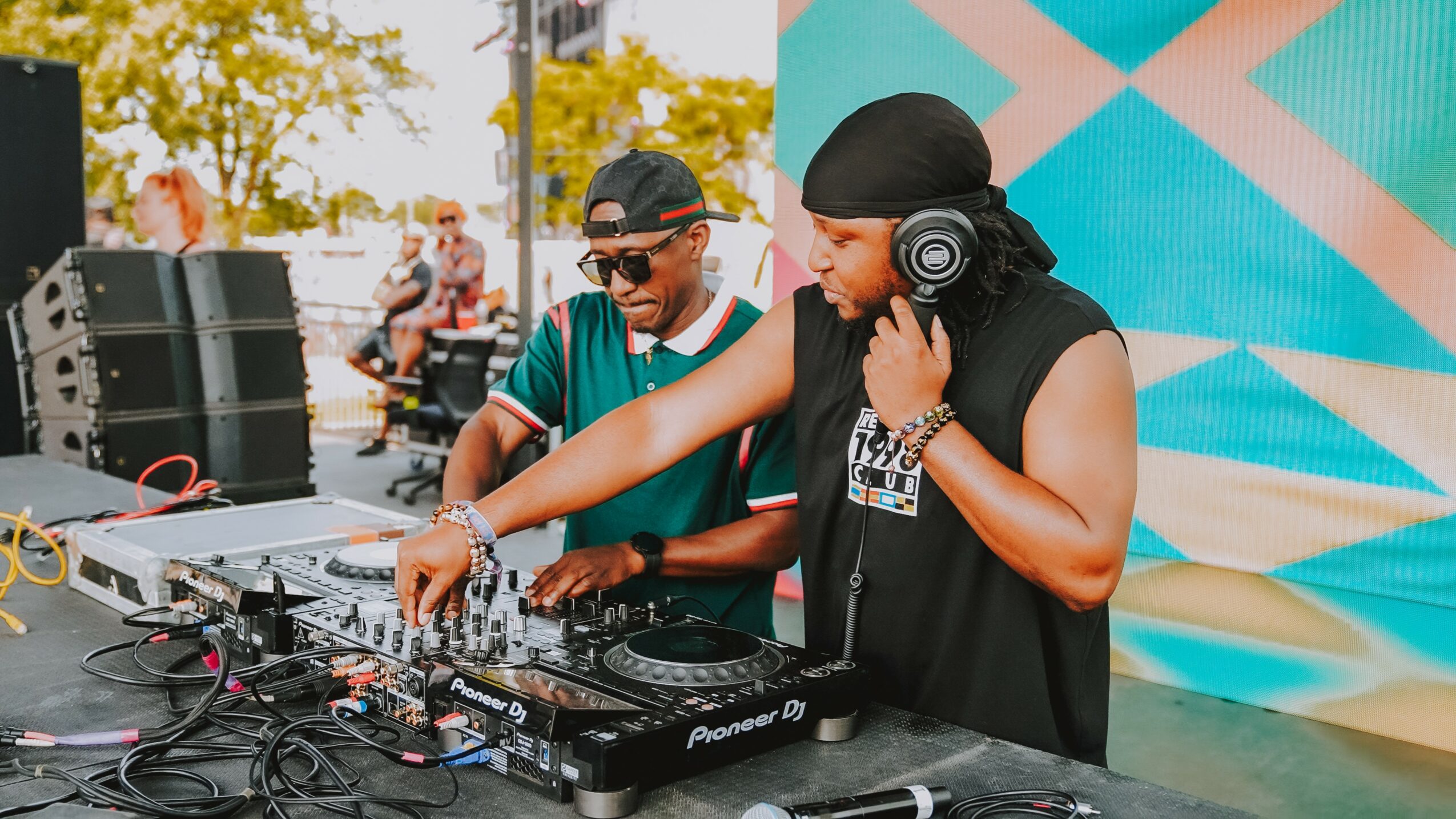 Two DJs perform outdoors with equipment, one wearing headphones and sunglasses, the other adjusting controls, colorful background behind them.