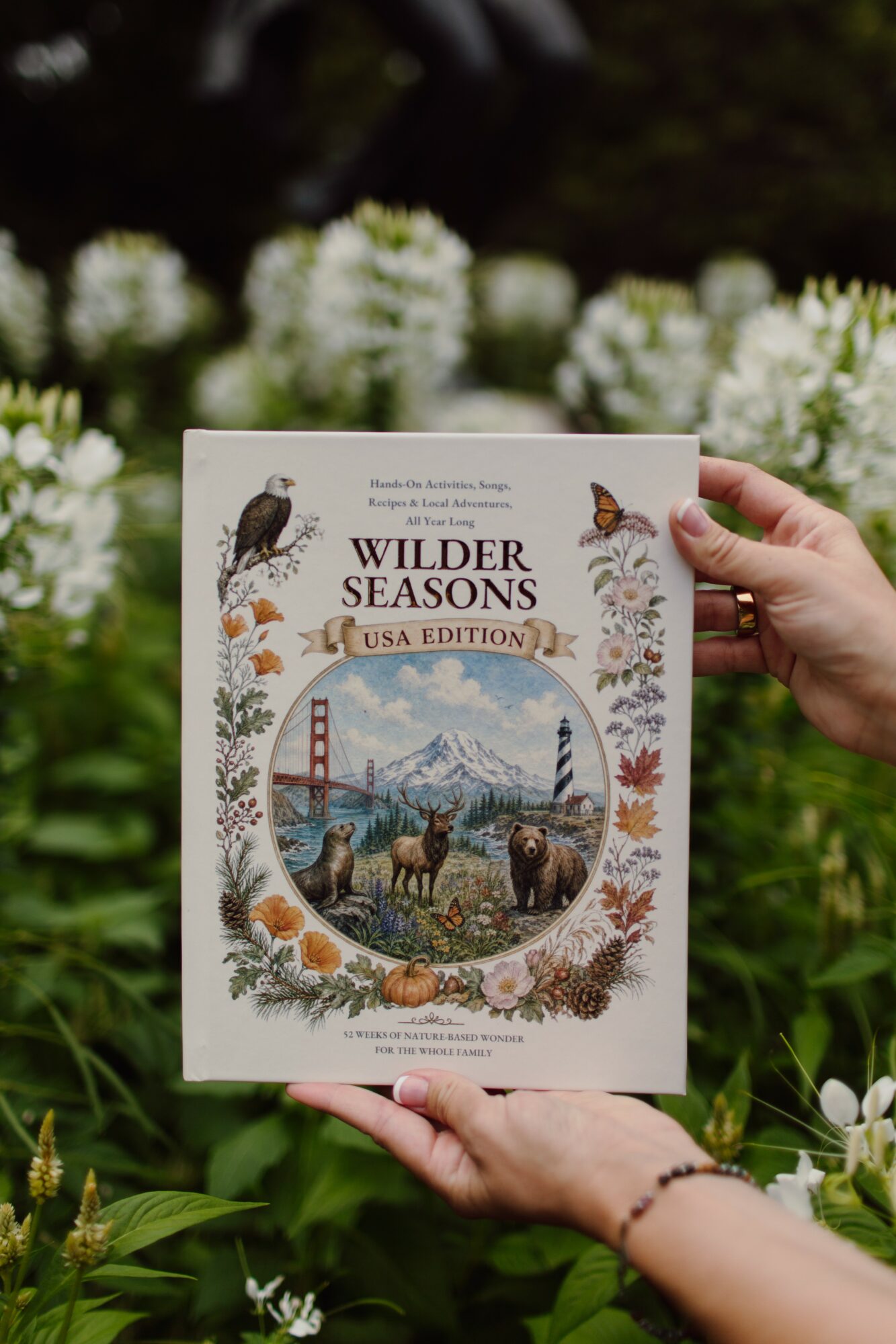 Person holding a book titled 'Wilder Seasons' in front of white flowers, with a nature scene on the cover.