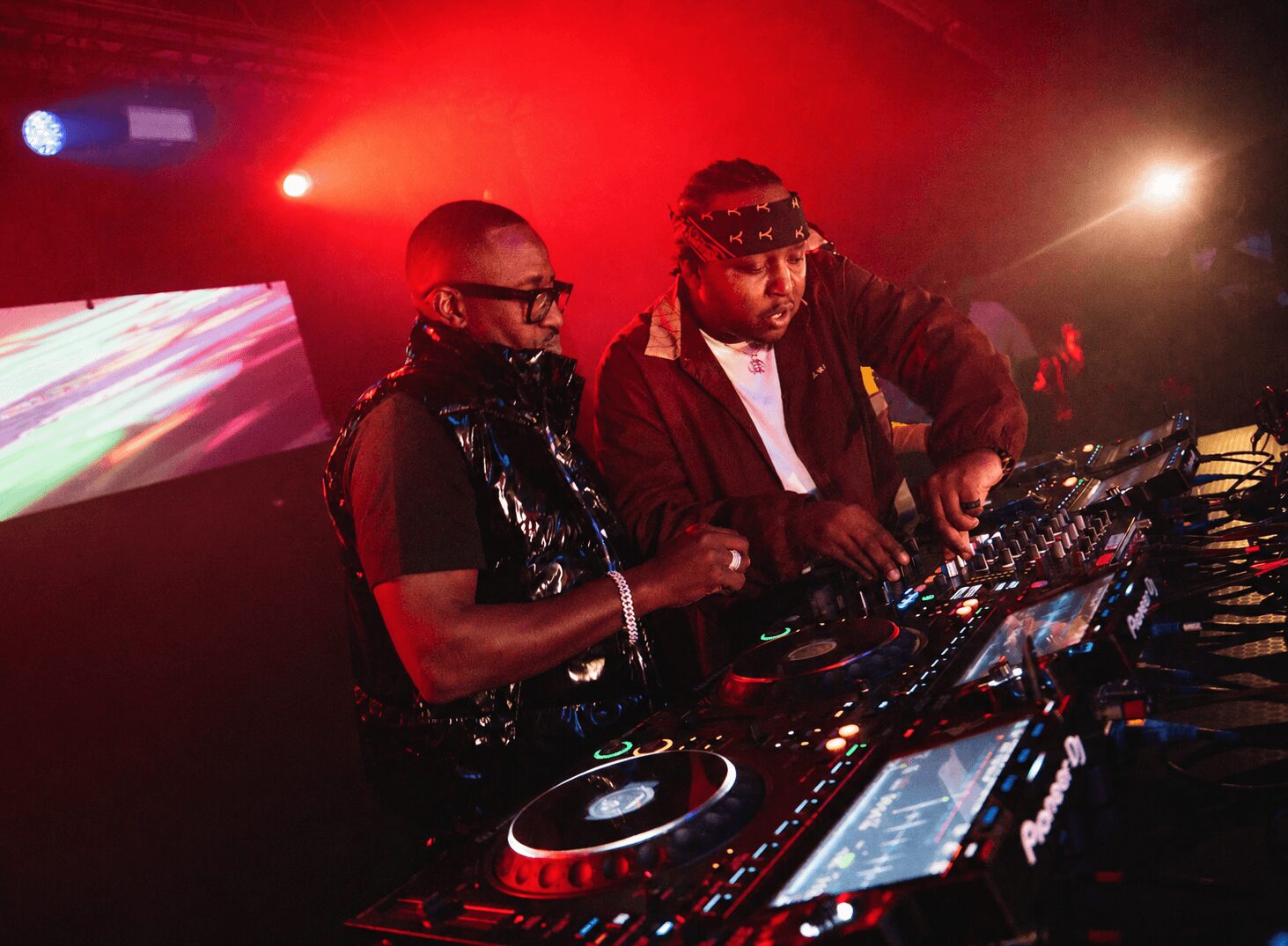 Two DJs perform at a nightclub with colorful lights and fog, using turntables and mixers.