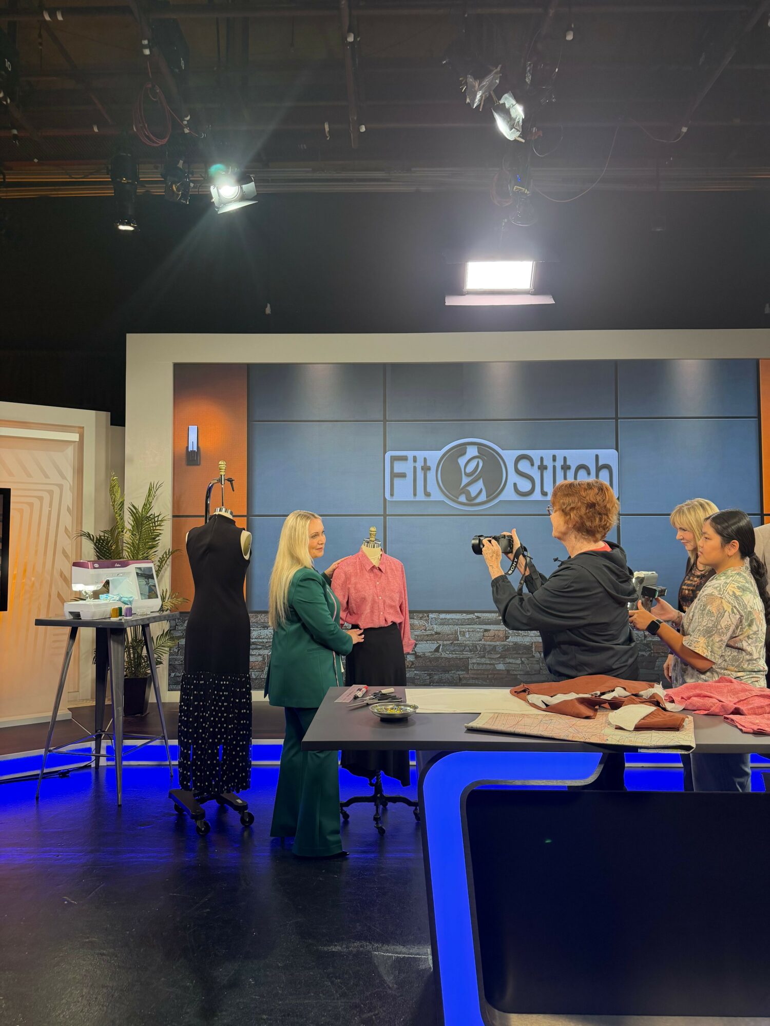 People sitting and standing in a studio with a large screen displaying a logo, some equipment, and a person speaking.