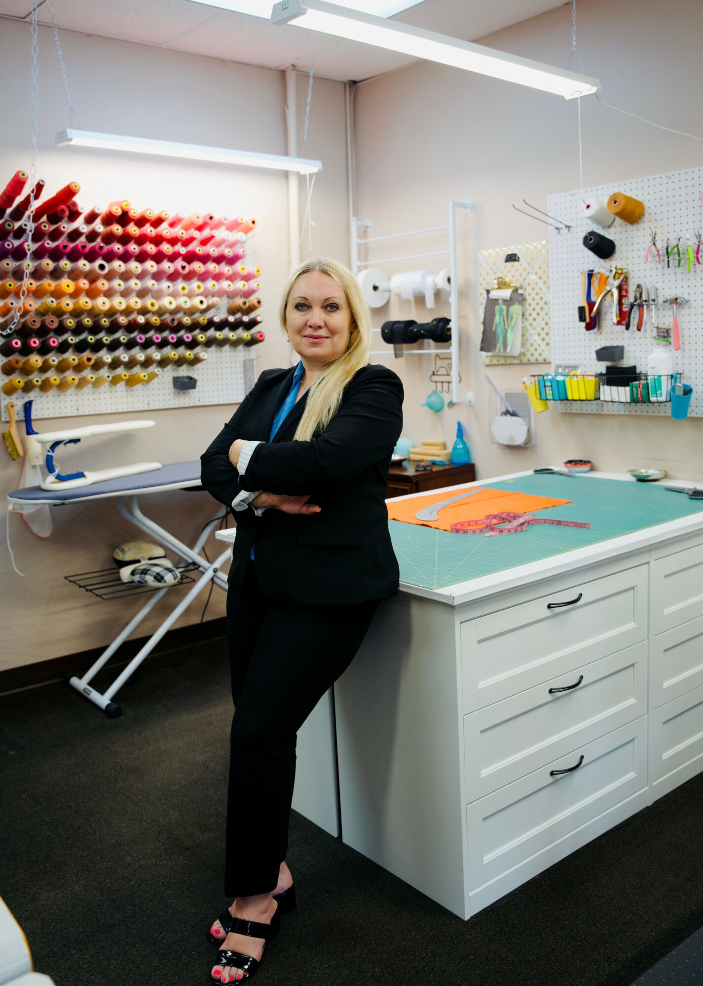 Woman standing in a sewing or craft room with colorful thread spools on wall, work table, and tools on wall, smiling.