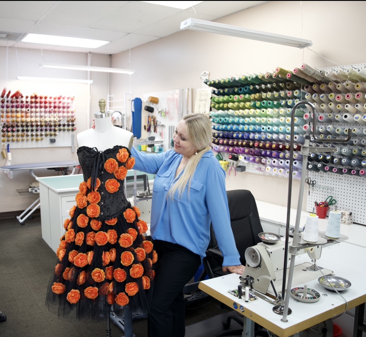 Woman working on a dress with orange flowers in a sewing studio, surrounded by thread spools and sewing tools.