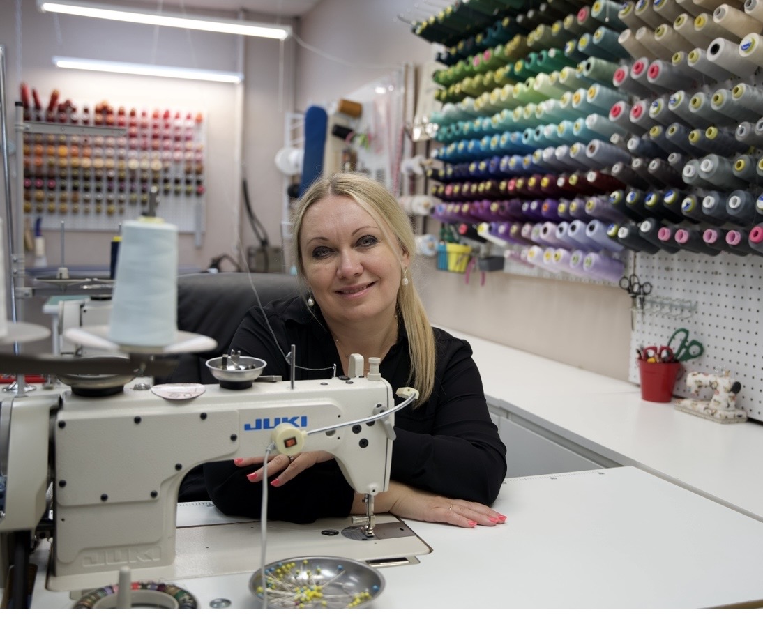 Woman with blonde hair smiling at sewing machine in fabric store, shelves with colorful spools of thread behind her.
