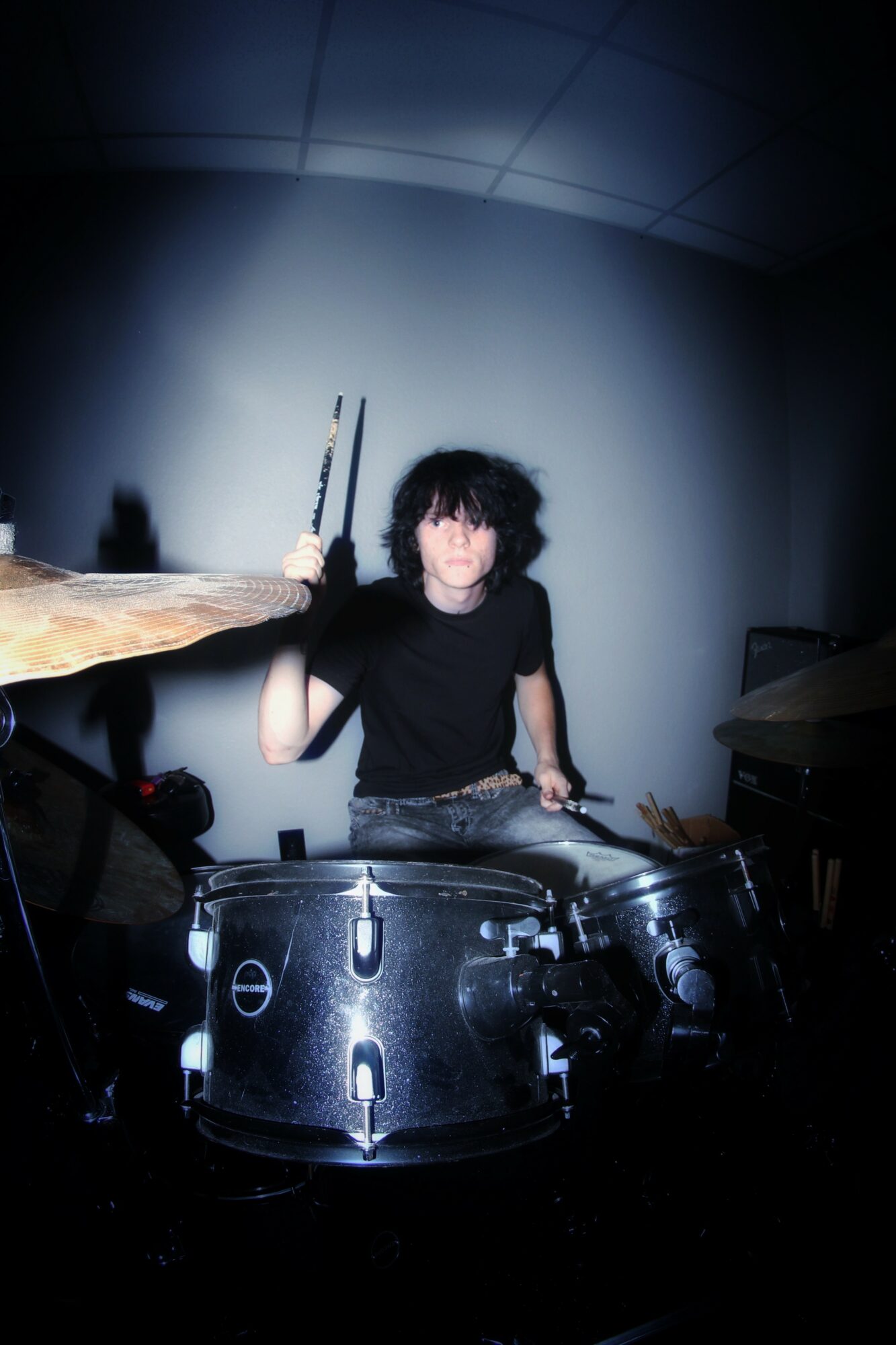 Person with curly hair playing drums in a dimly lit room, holding drumsticks, with a dark background.