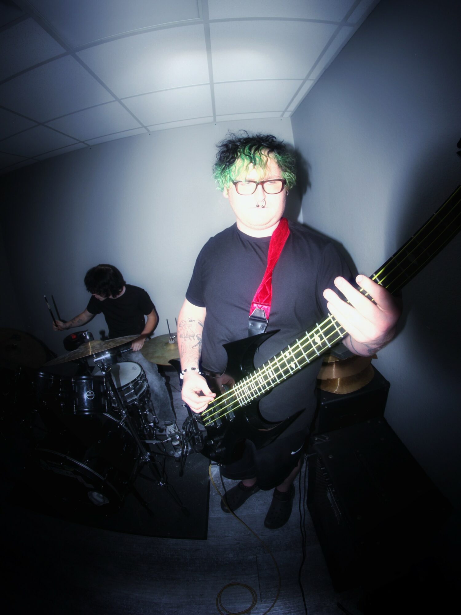 Person with green hair and glasses playing bass guitar in a small room, with another person playing drums in the background.
