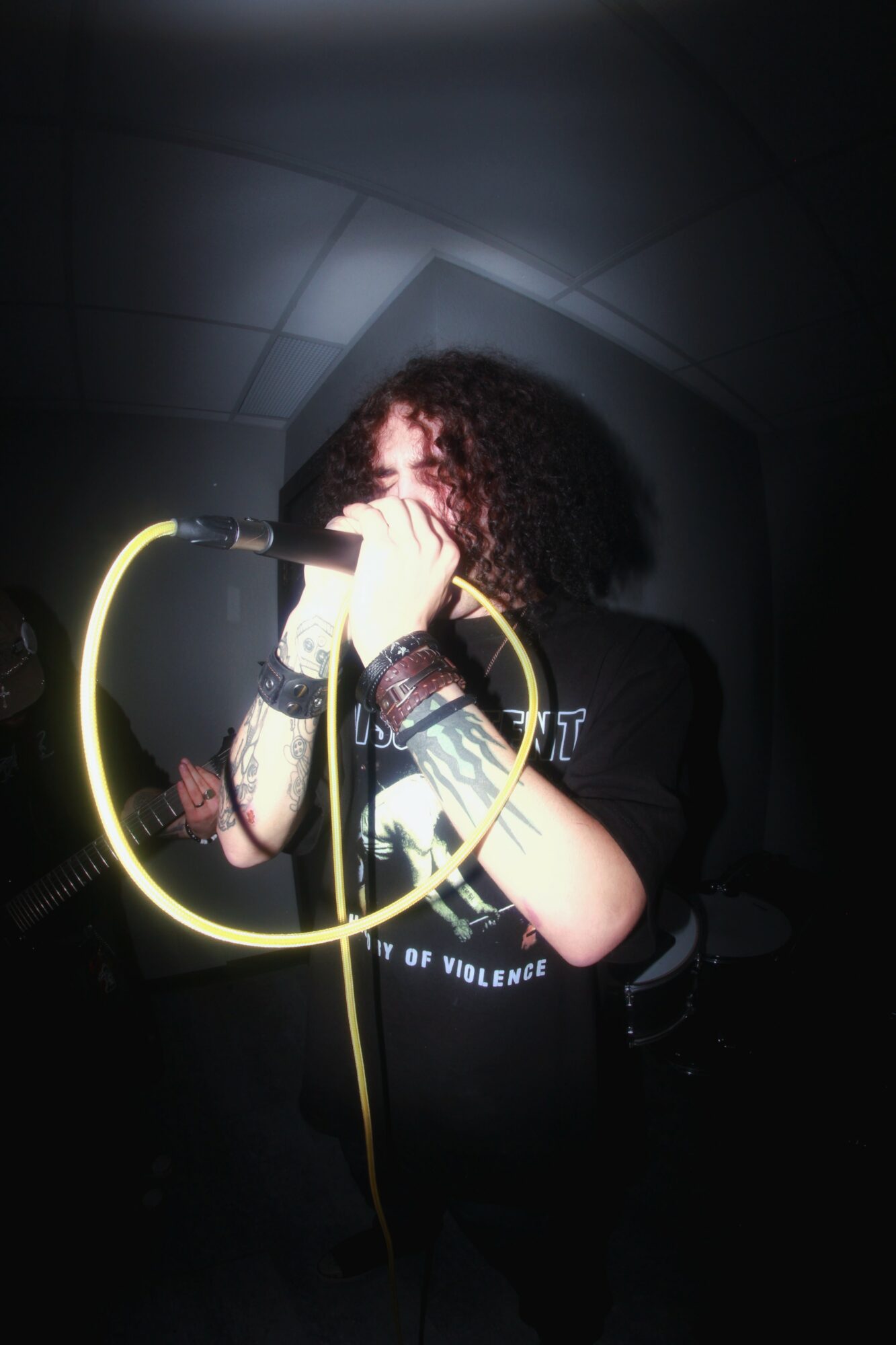 Person with curly hair singing into a microphone, holding a yellow cable, in a dark setting.