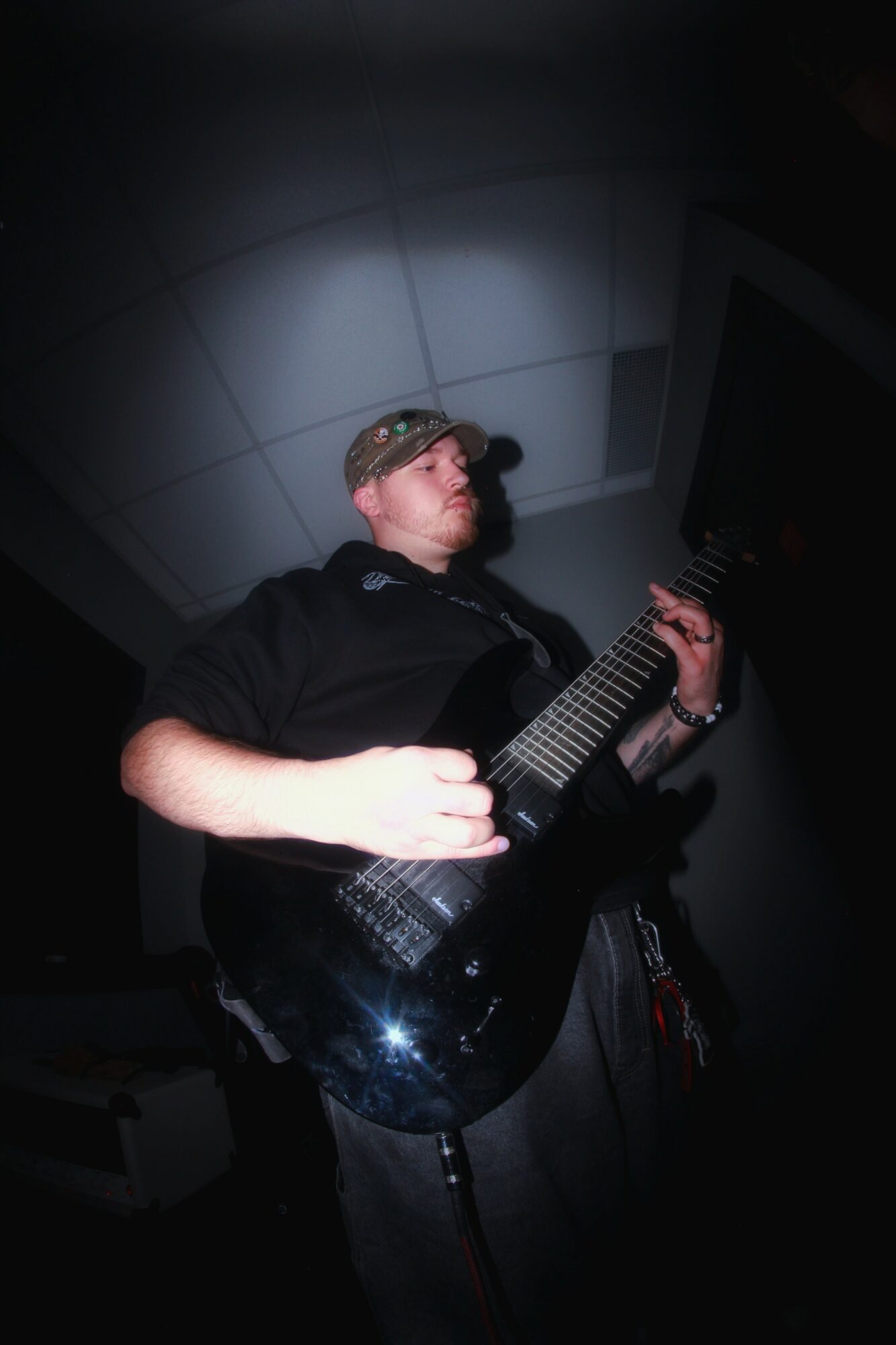 Person playing an electric guitar in a dark room with a tiled ceiling, illuminated by a spotlight.
