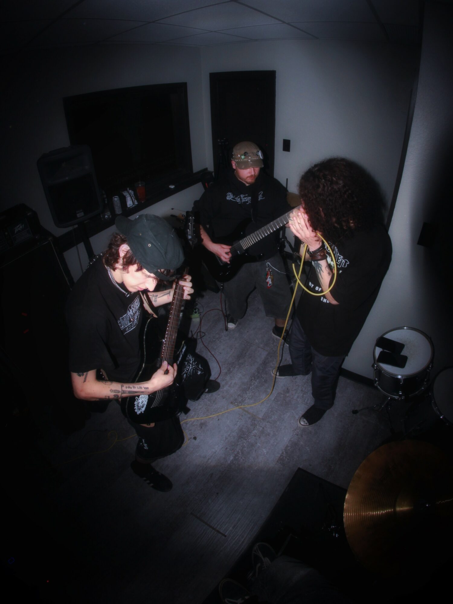 Three people in a small room, two playing guitars and one singing into a microphone, with music equipment around.