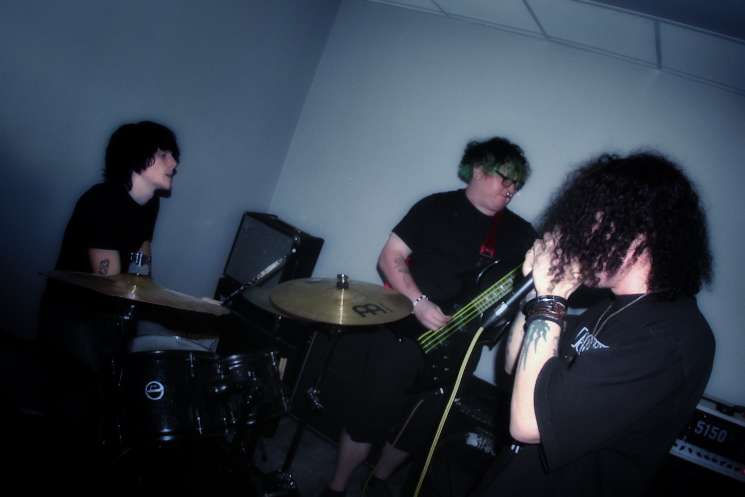 Three musicians perform in a dimly lit room, one playing drums, one with a bass guitar, and one singing into a microphone.