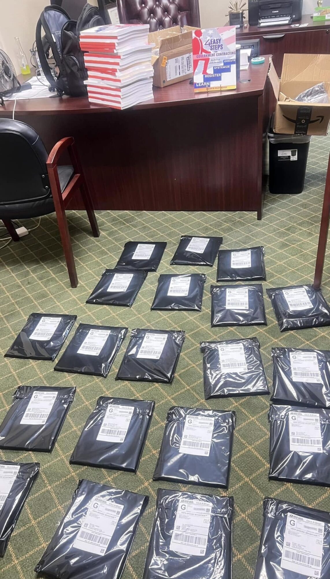 Multiple sealed packages on a green carpeted floor in front of a desk with stacks of books and office supplies.