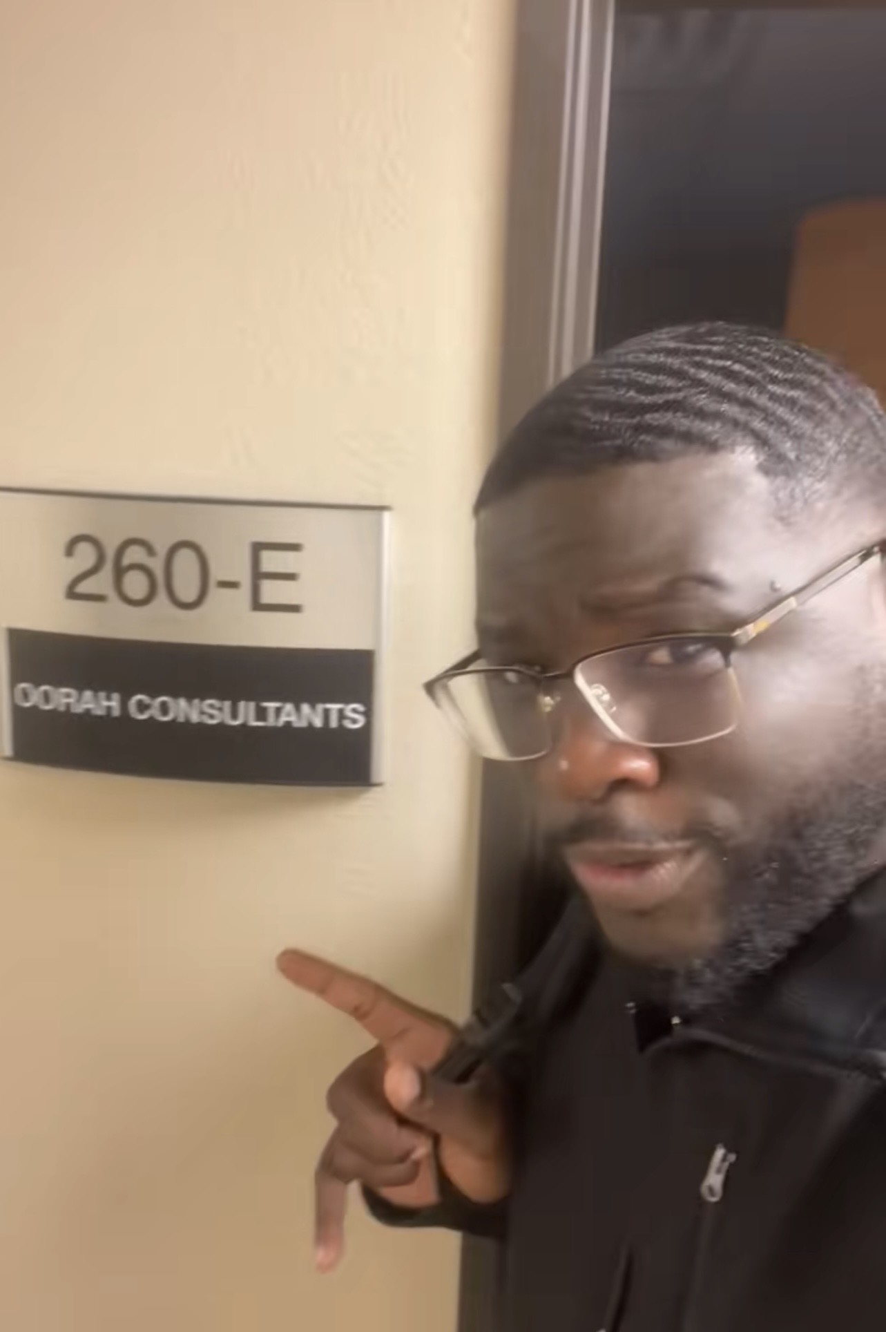 Man with glasses pointing at a room number sign on a wall, with a window nearby.