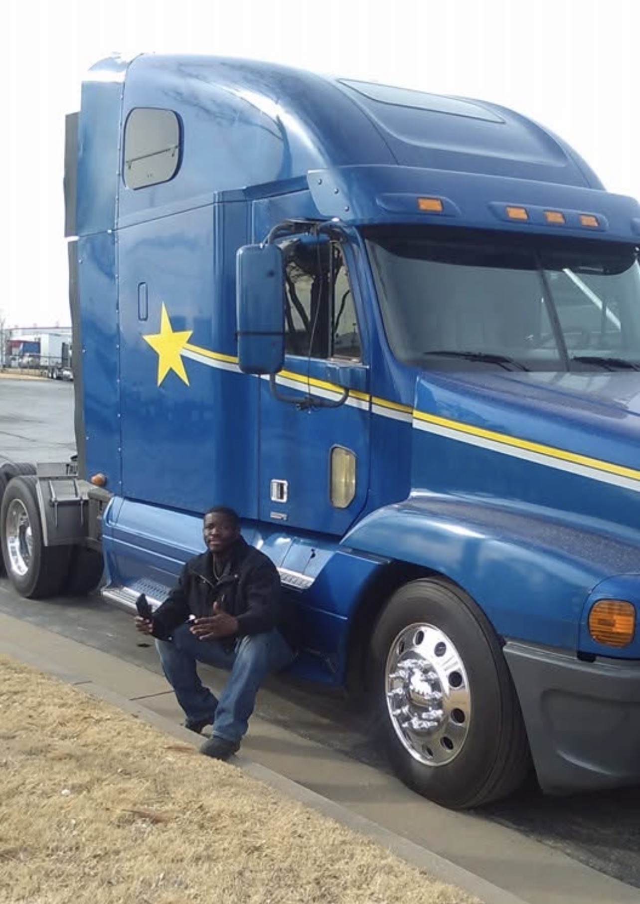 Person sitting on the ground in front of a large blue semi-truck with a yellow star and stripe, parked on a street.