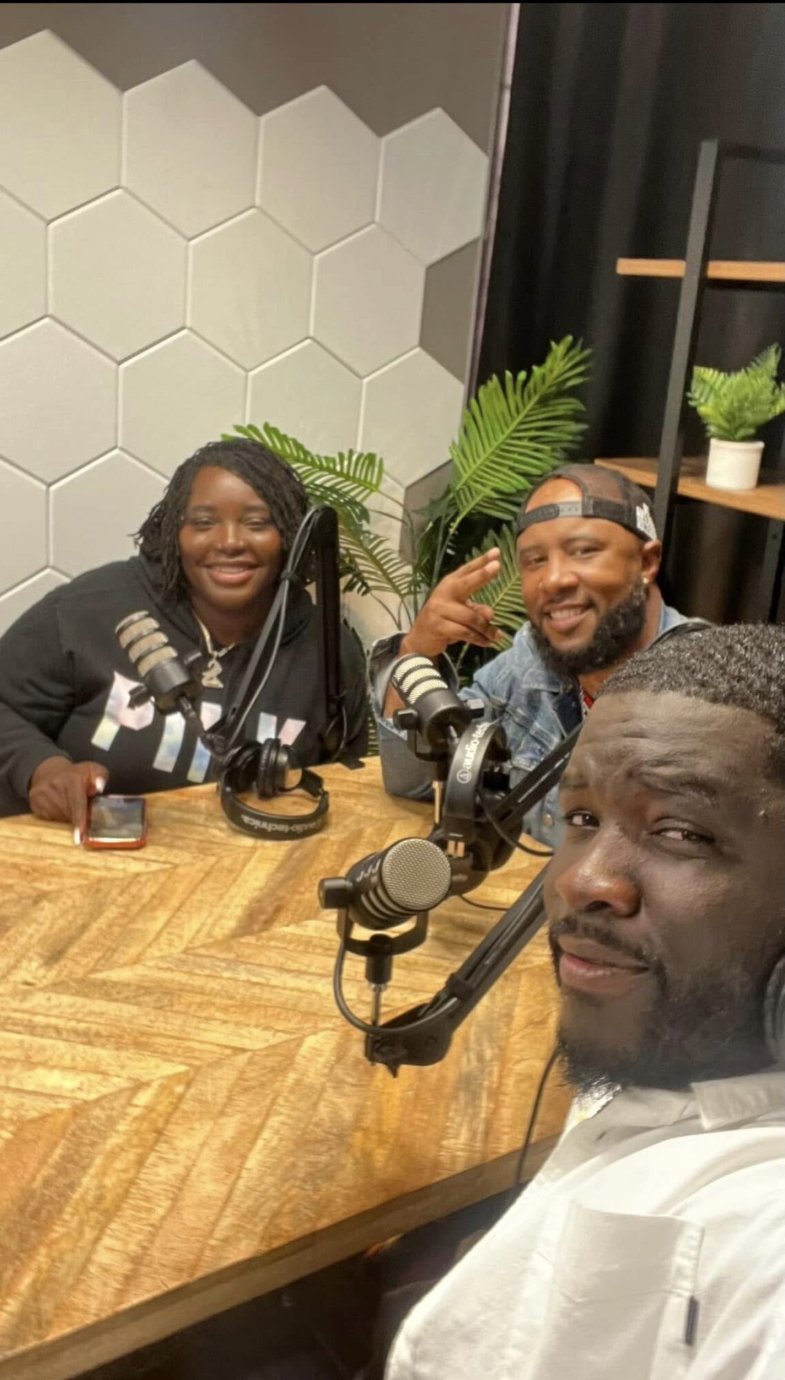 Three people sitting at a wooden table with microphones, smiling, in a room with hexagonal wall tiles and plants.