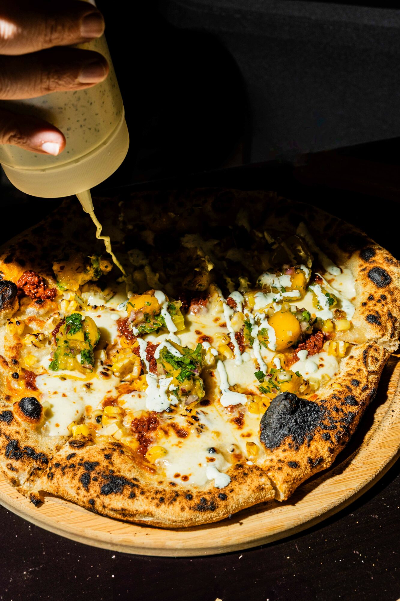 Pizza with eggs, herbs, and cheese on a wooden board, with a hand squeezing sauce on top.