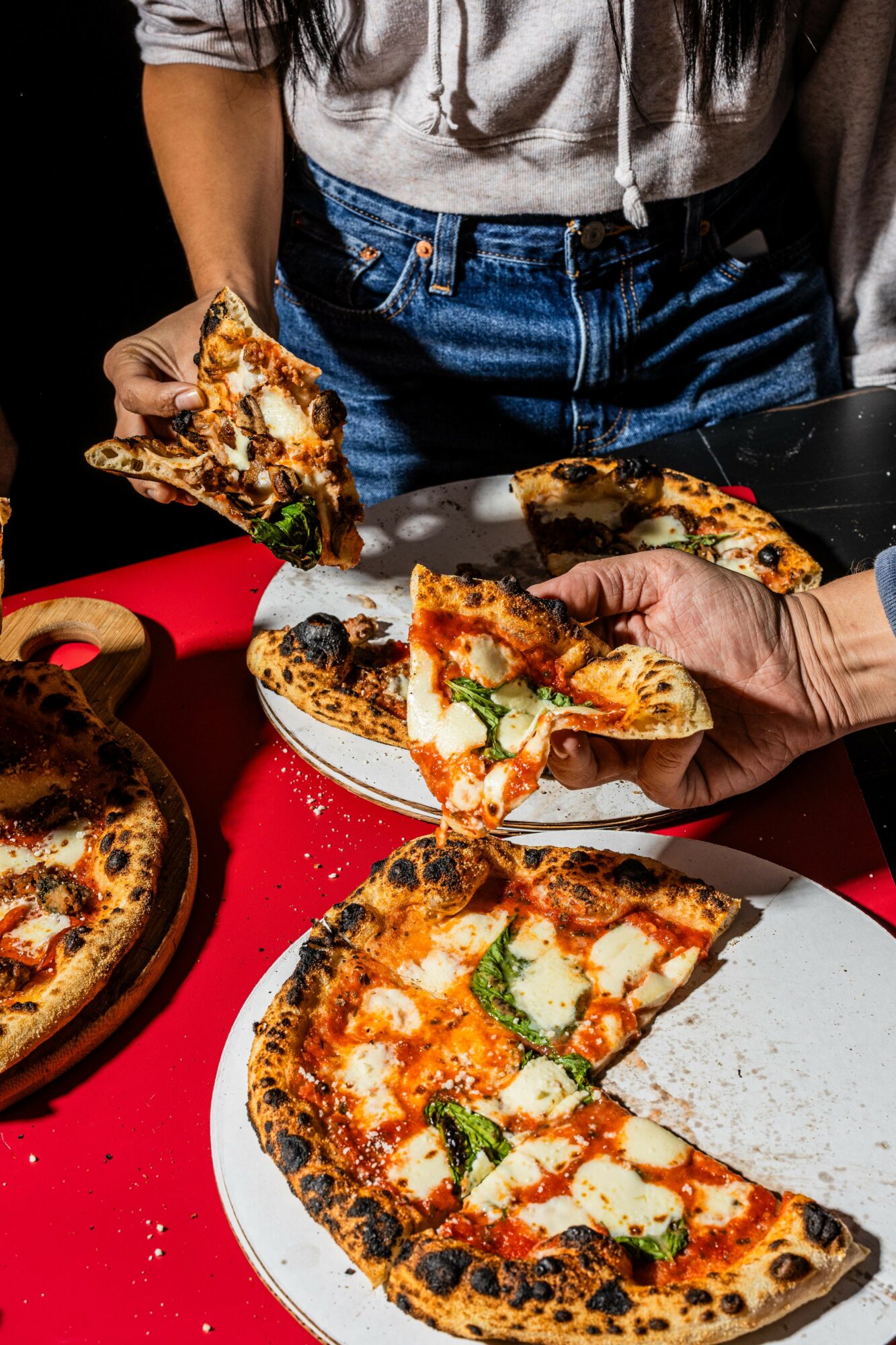 Person serving pizza with slices on plates, pizza topped with cheese, tomatoes, and basil, on a red table.