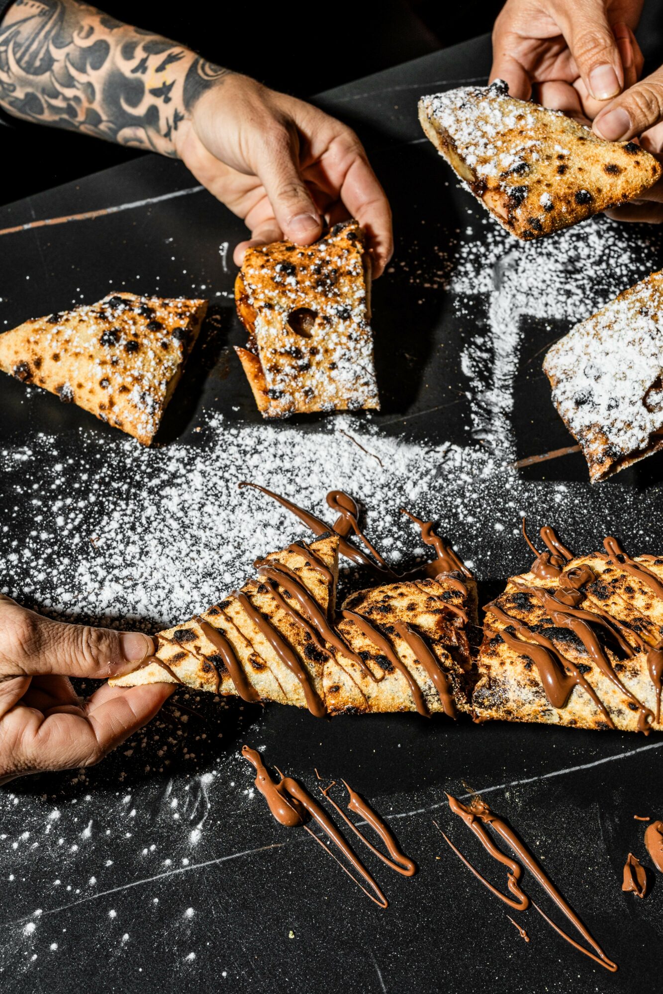 Hands reach for slices of cake and a pastry on a black plate, with chocolate drizzle and powdered sugar, on a dark surface.