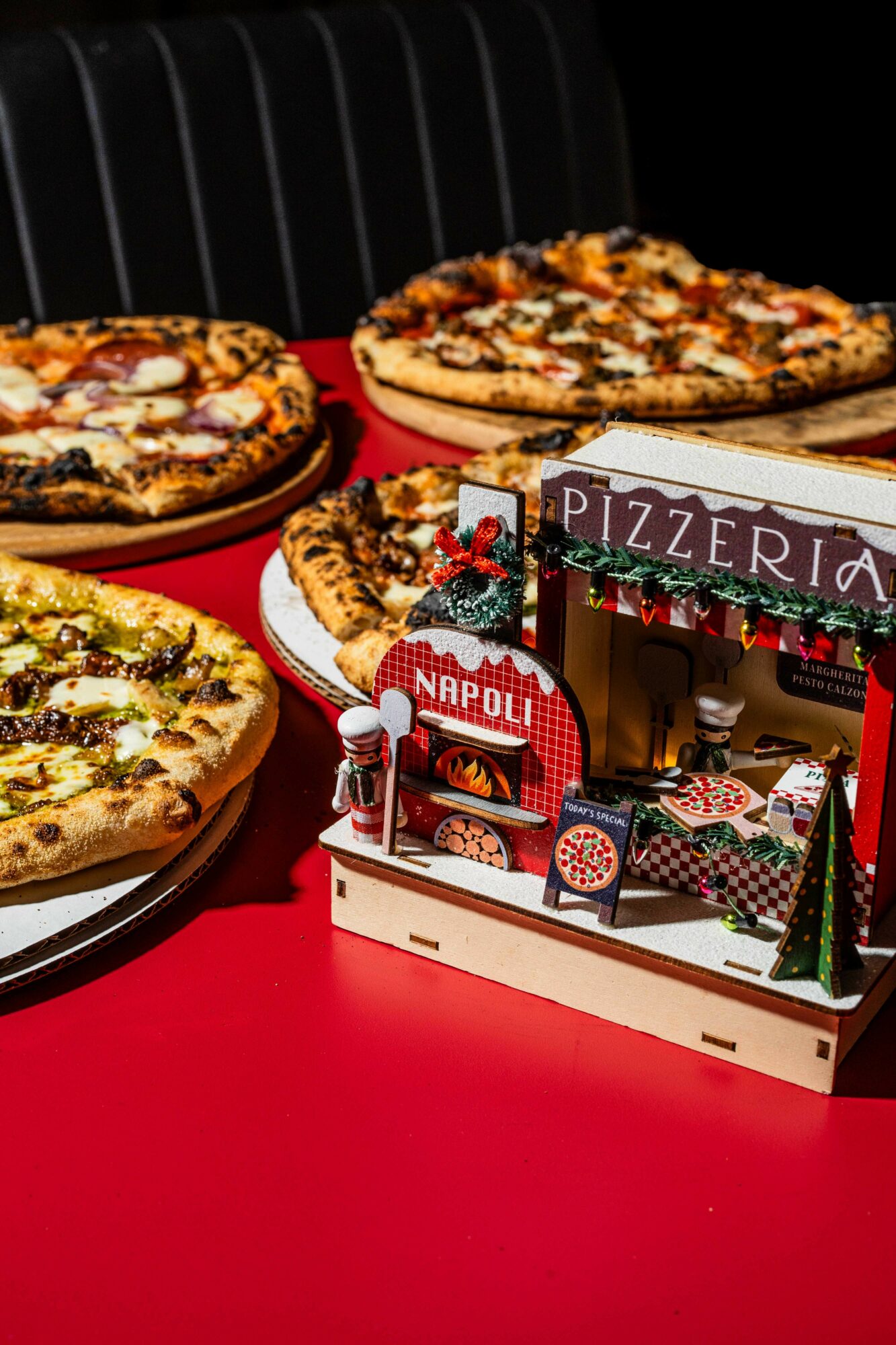 Three pizzas on a table with a miniature pizzeria display in front, set on a red surface against a dark background.