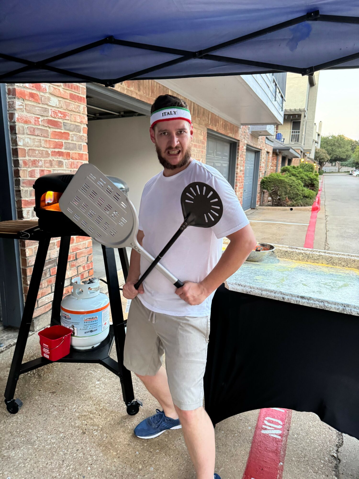 Person grilling outdoors, holding a spatula, wearing a white shirt and shorts, standing next to a barbecue grill.