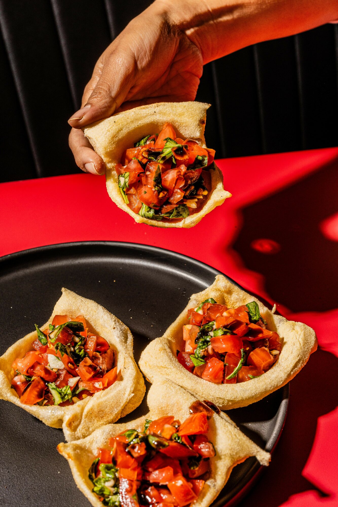 Hand holding a taco above a black plate with four tacos filled with vegetables and sauce, red background.