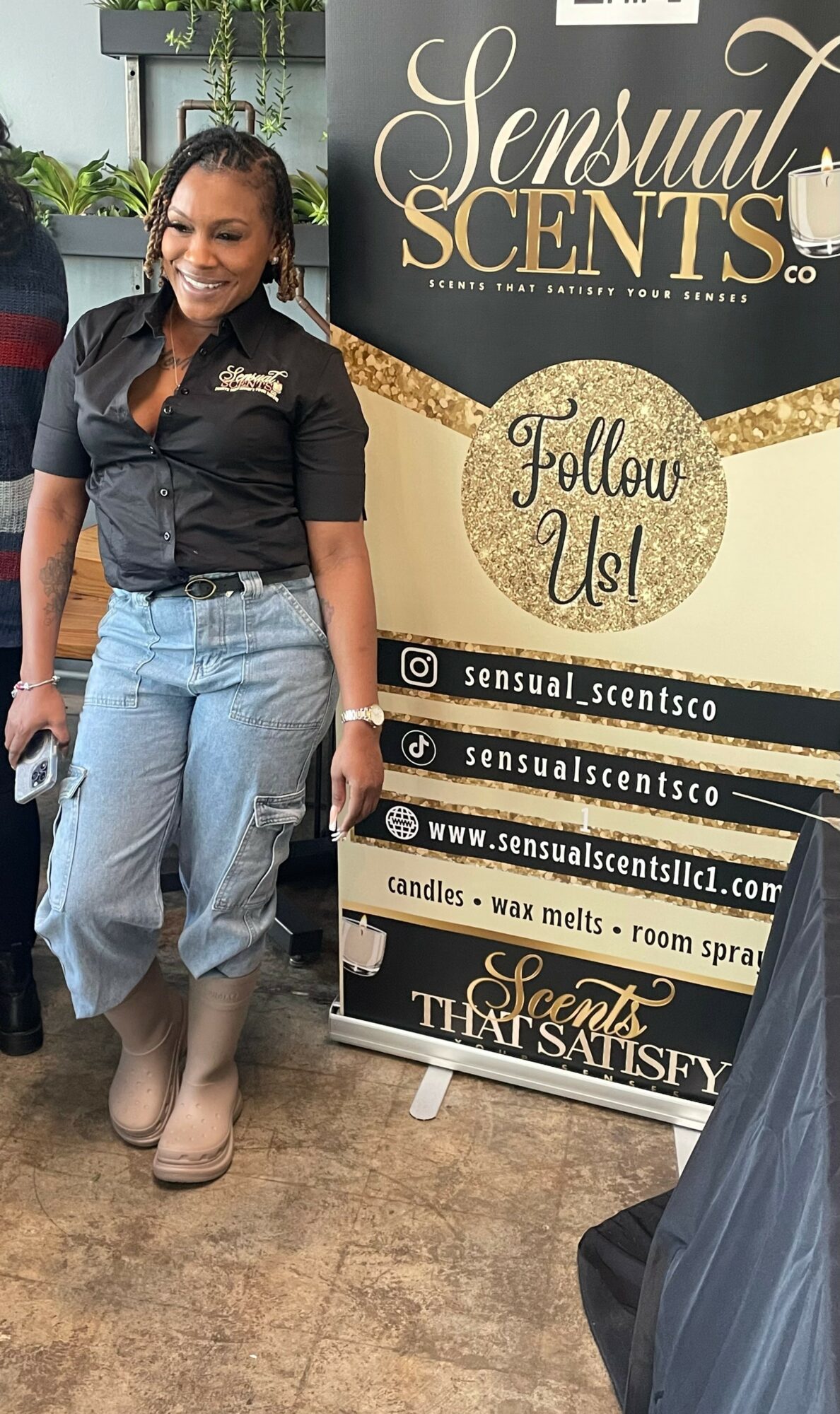 Smiling woman standing next to a promotional sign for Sensual Scents, wearing a black shirt and light blue jeans.