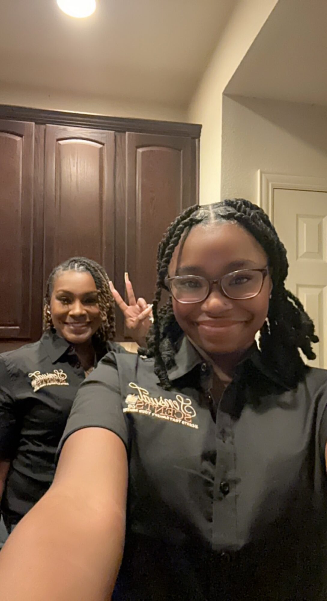 Two women smiling and taking a selfie indoors, with dark wooden cabinets in the background.