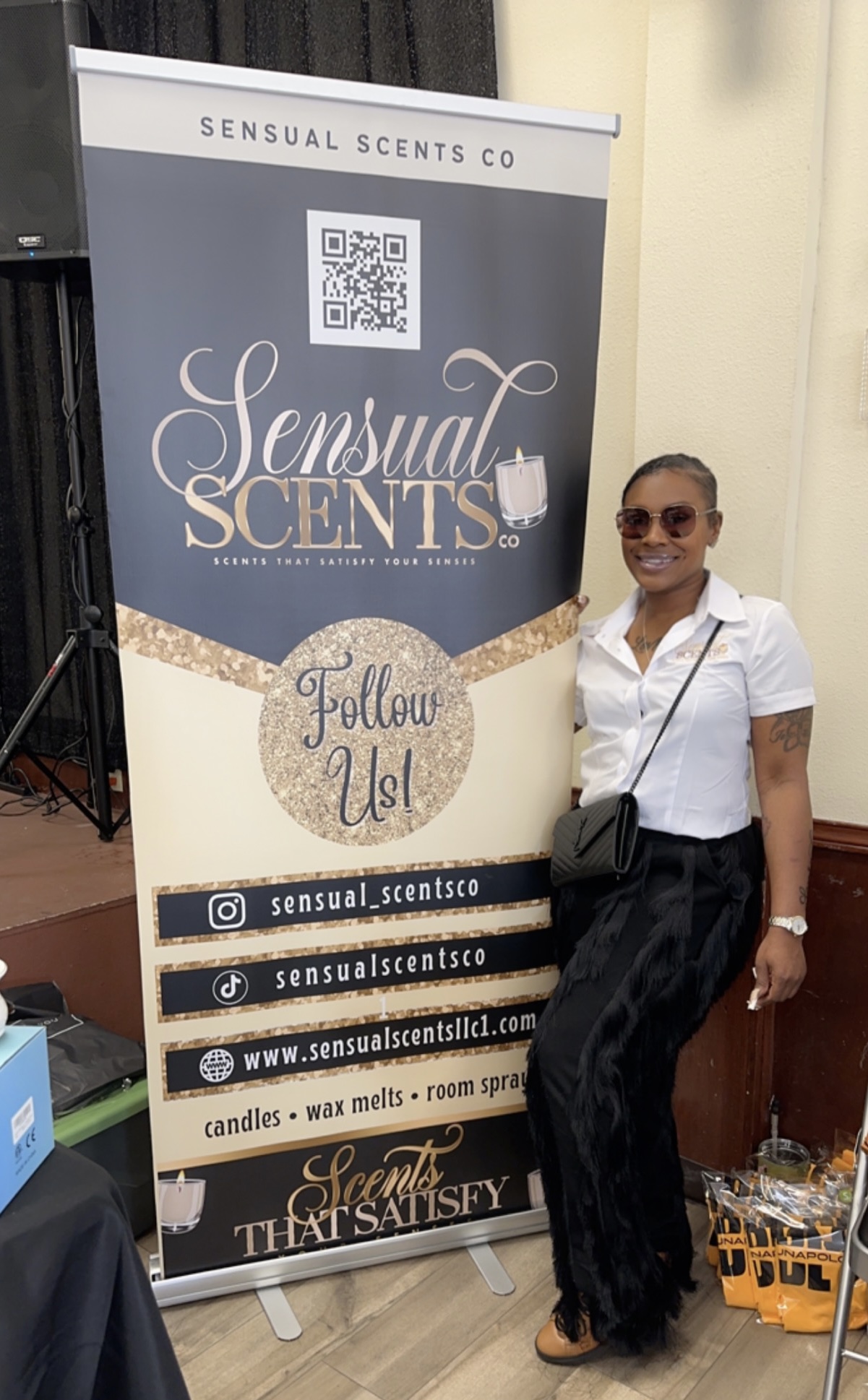 Woman with sunglasses and white shirt standing next to a Sensual Scentsco banner, smiling outdoors.