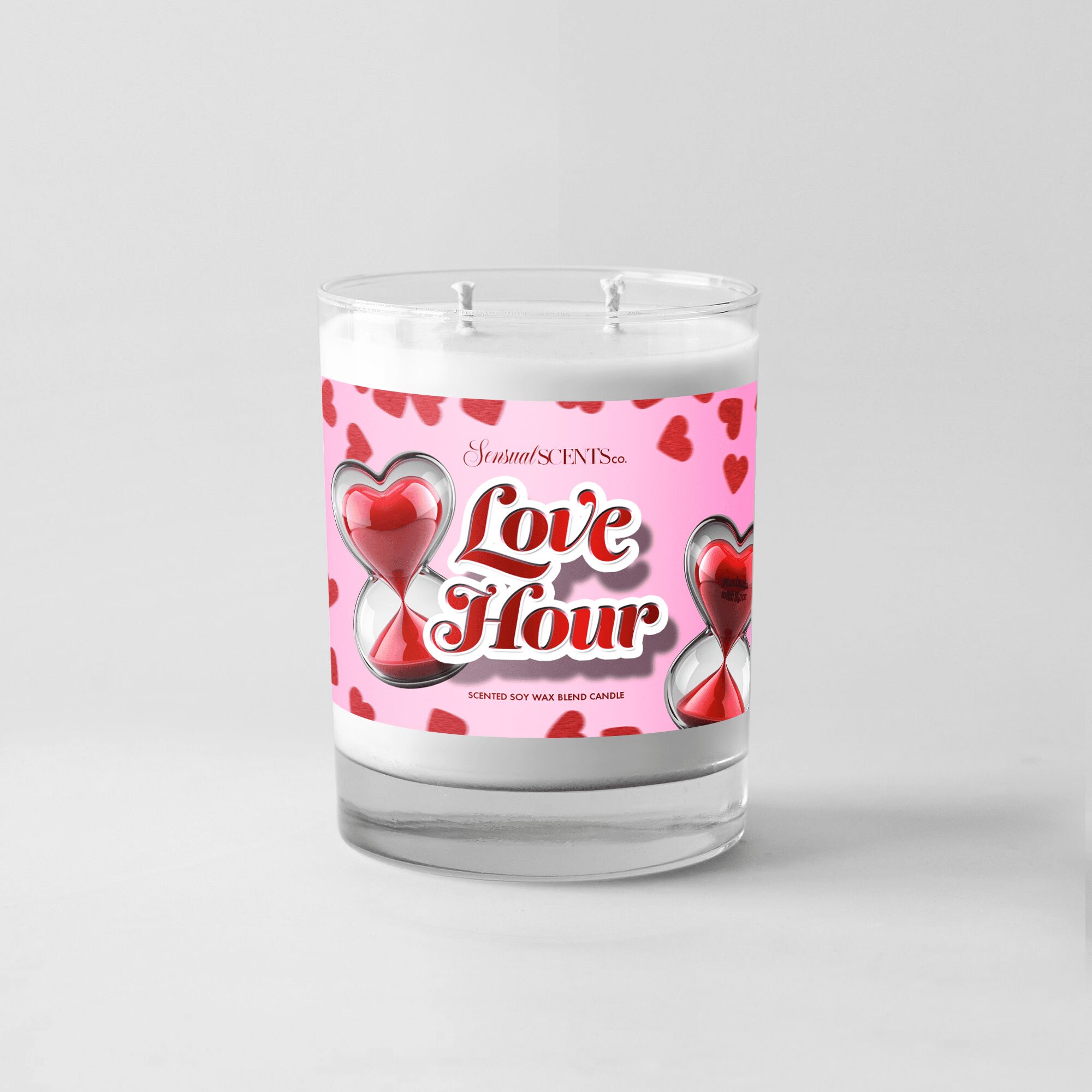 A white candle in a glass container with a pink label featuring hearts and an hourglass, labeled 'Love Hour'.