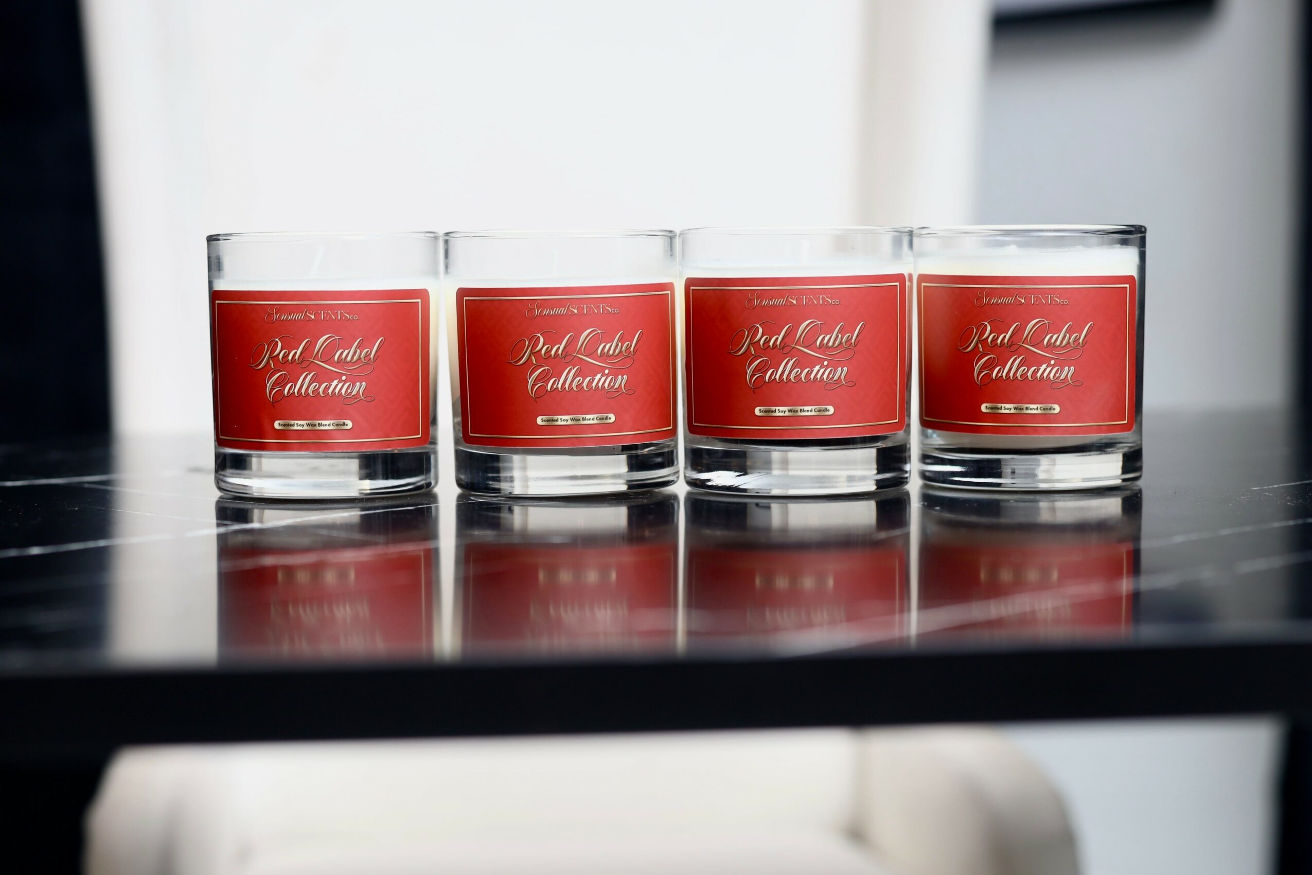 Four candles with red labels on a black surface, reflected below, with a blurred background.
