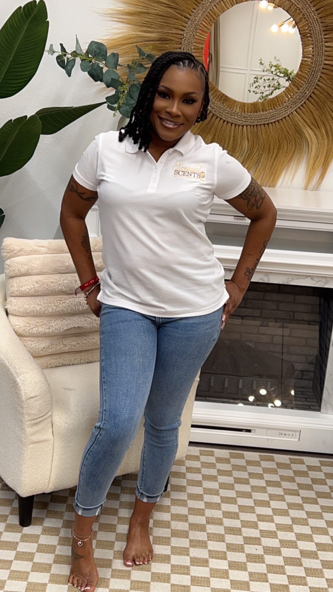 Woman with braided hair in a white polo shirt and jeans standing indoors, smiling, with a mirror and decorative wall behind her.