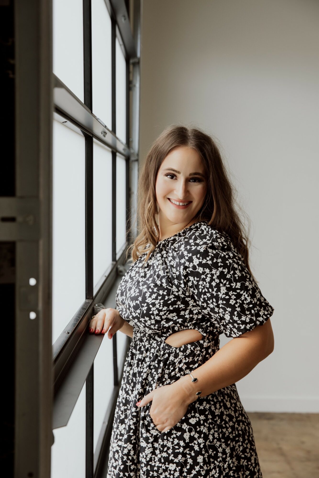 Young woman with long hair smiling, standing near a large window with black frames, wearing a black and white patterned dress.