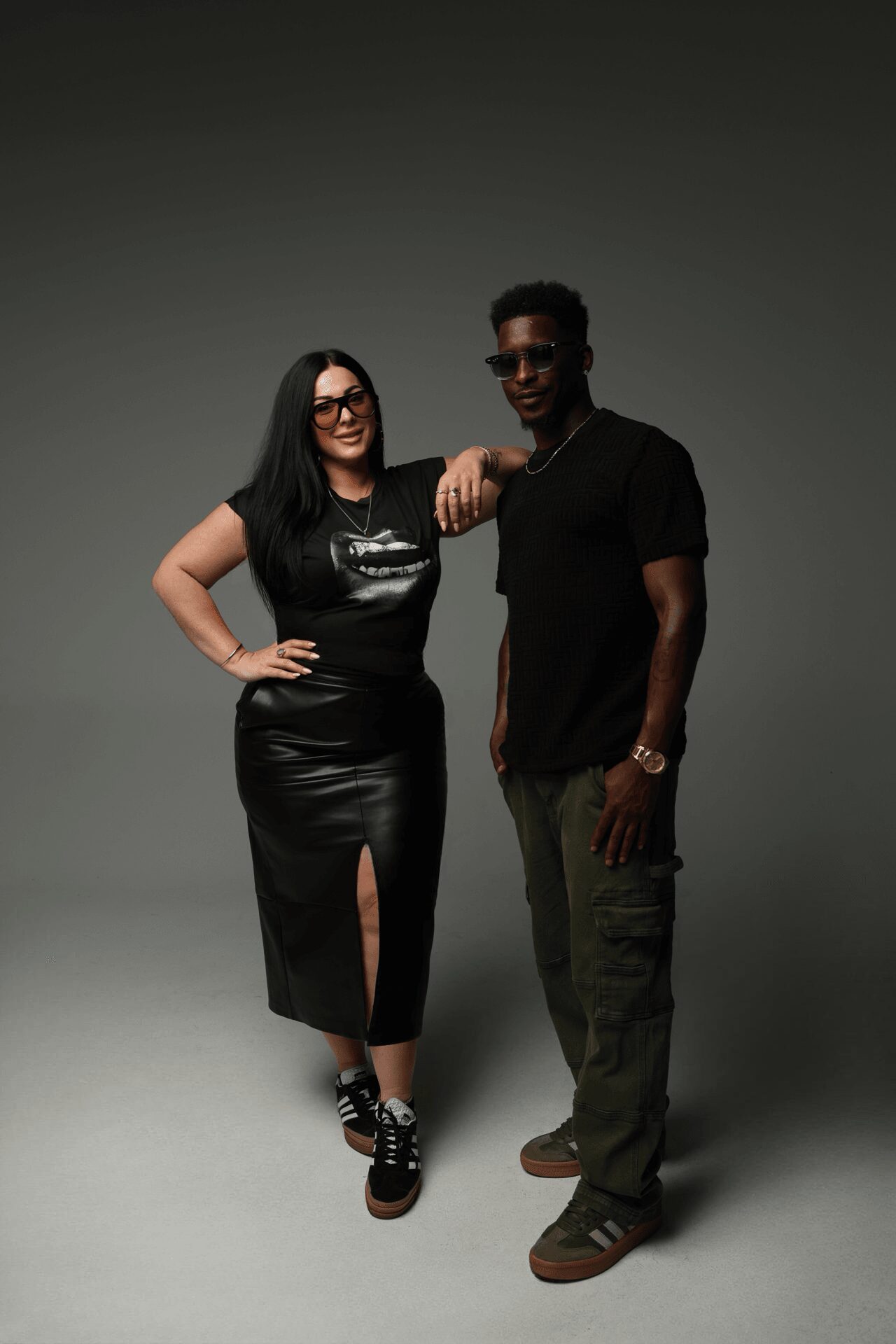 Two people stand together, one with long dark hair and glasses, the other with short curly hair, both dressed casually.