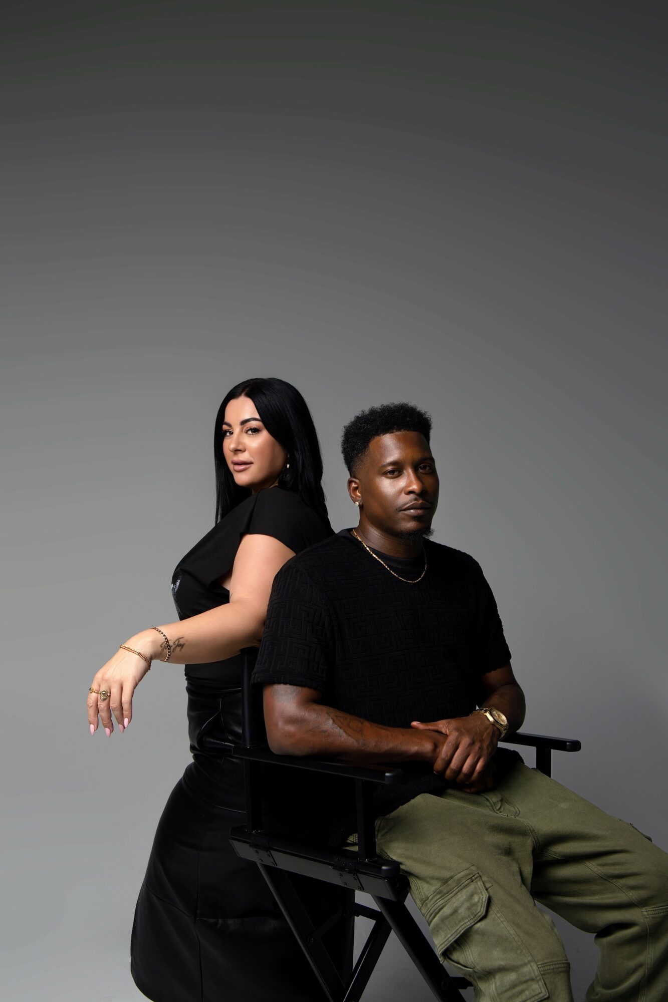 Two people sit against a plain gray background, one woman and one man, both looking at the camera.