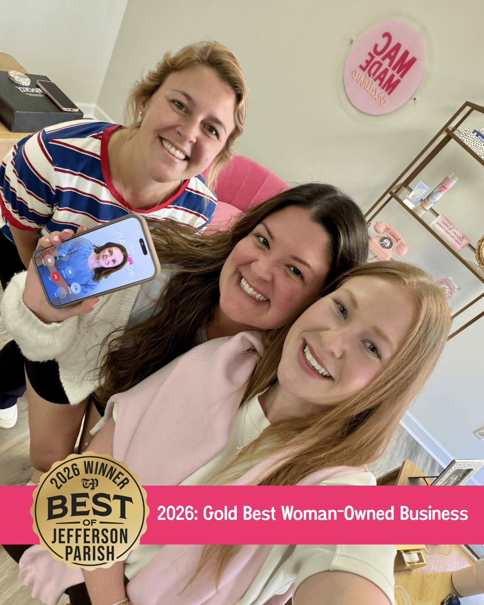 Three women smiling and taking a selfie in a salon or beauty shop, with a pink sign and shelves in the background.