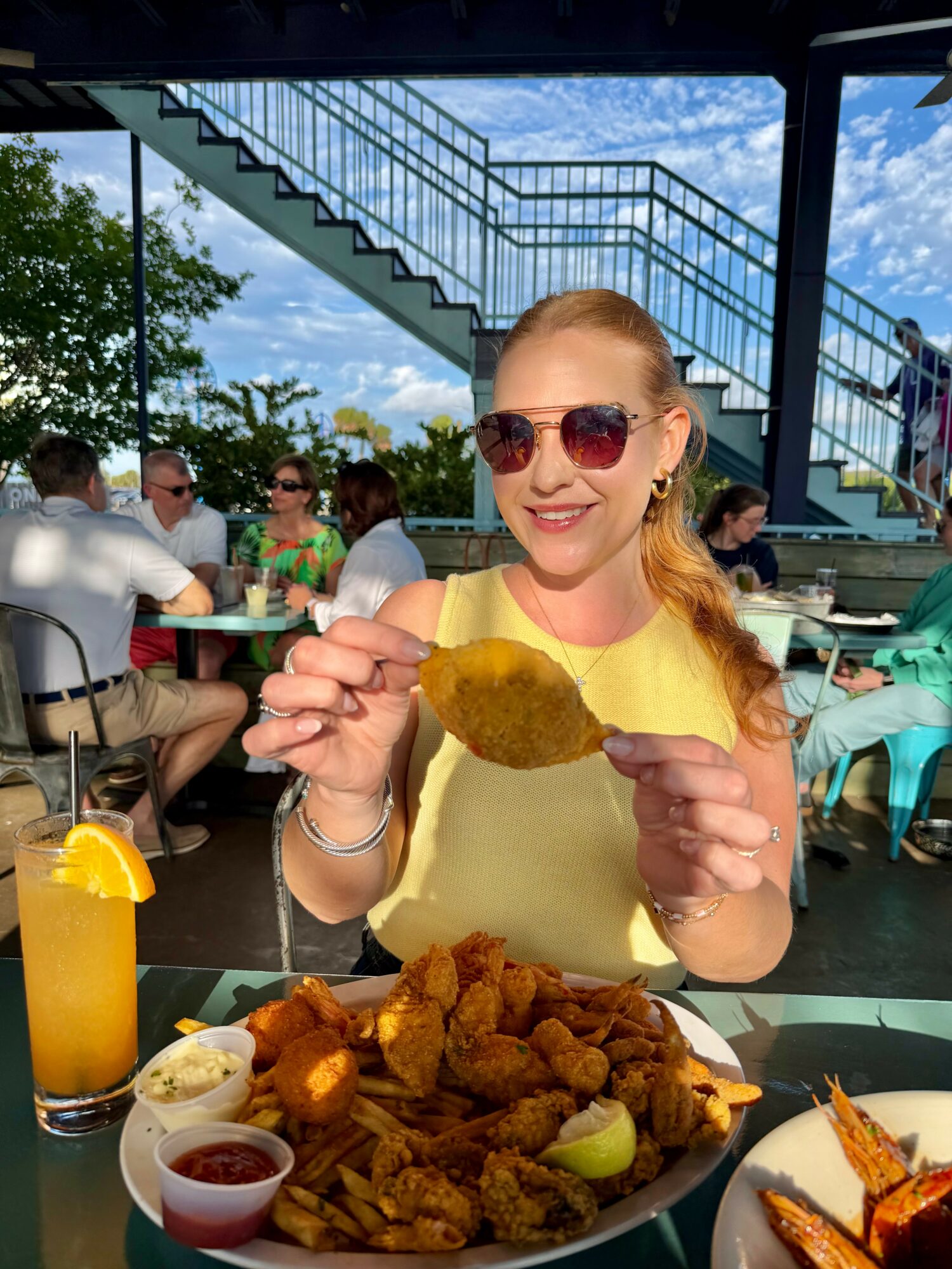 Woman with sunglasses smiling, holding fried chicken, seated at table with fried food and drinks outdoors.