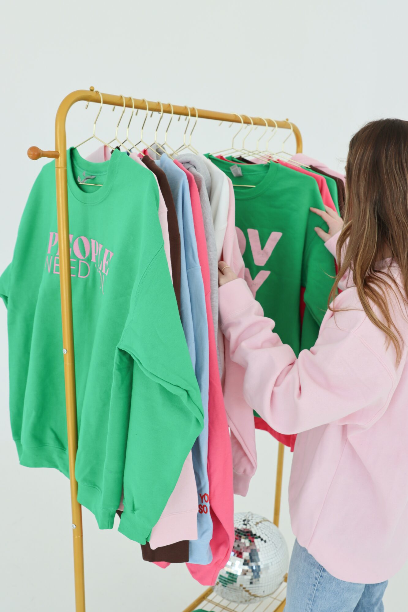 Person shopping for colorful sweatshirts on a clothing rack, wearing a pink jacket, with a soccer ball nearby.