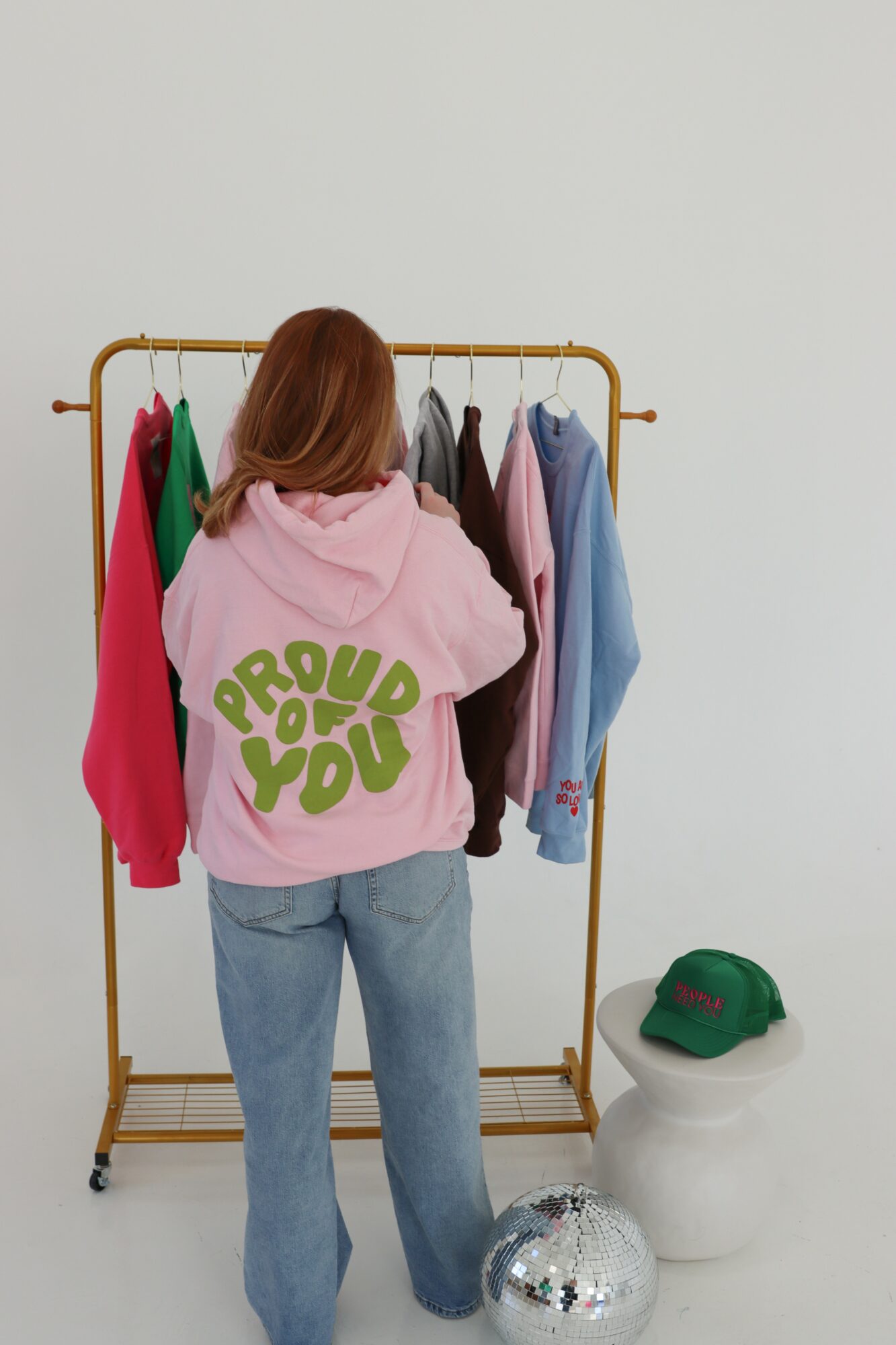 Person with red hair wearing a pink hoodie with green text, facing a clothing rack with various garments, against a plain white wall.