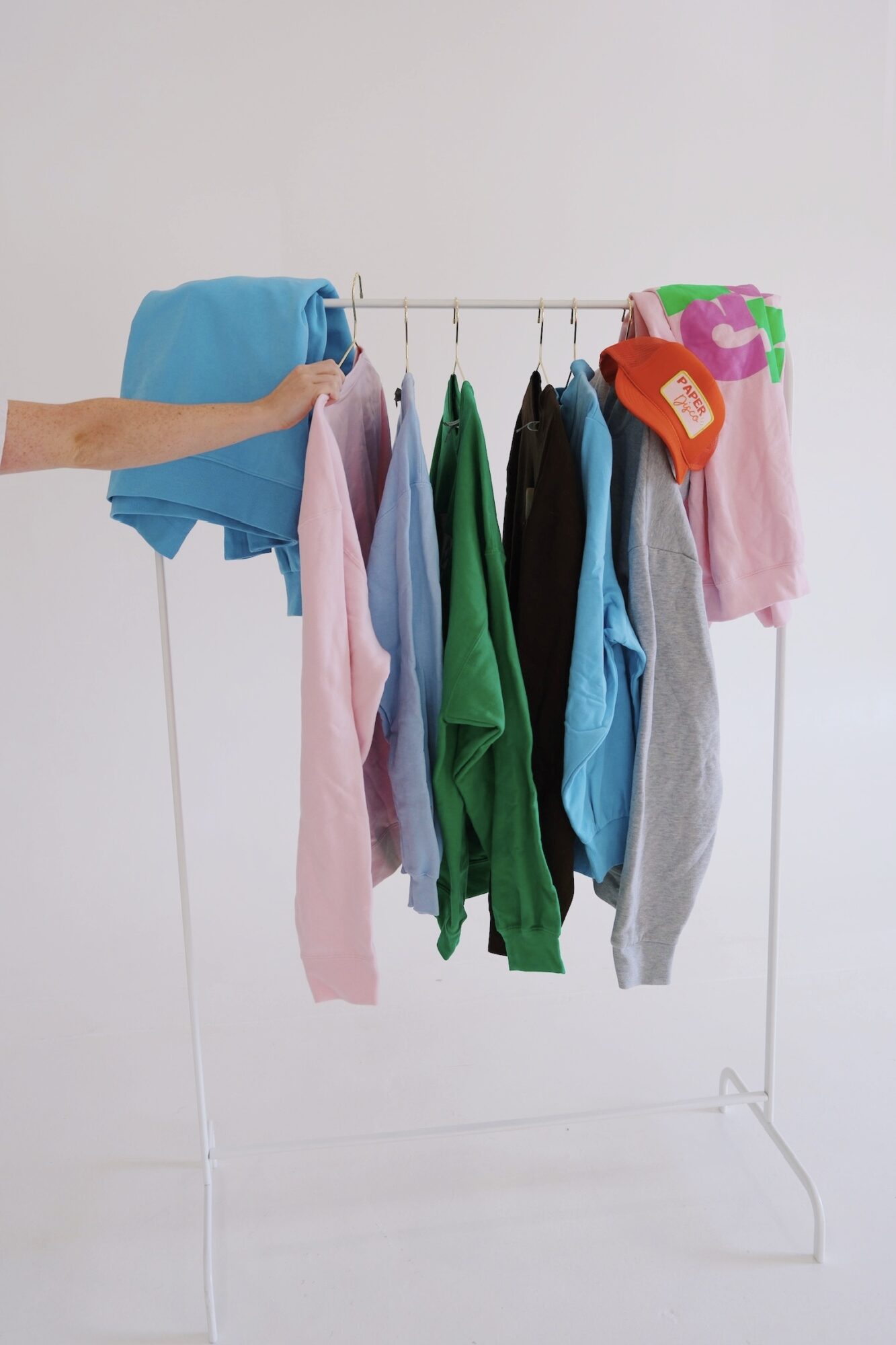 Clothes hanging on a rack with a hand holding a blue shirt, against a plain white background.