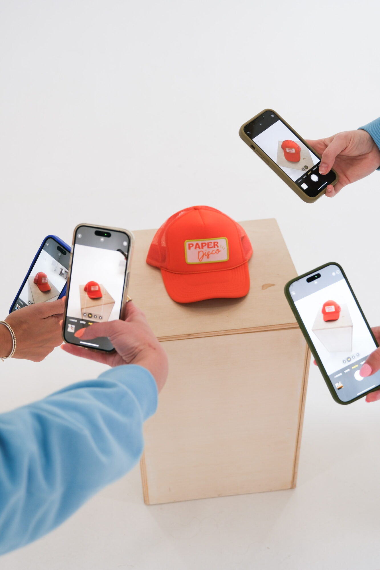 Three people take photos of a red cap with a paper label, on a wooden box, against a white background.