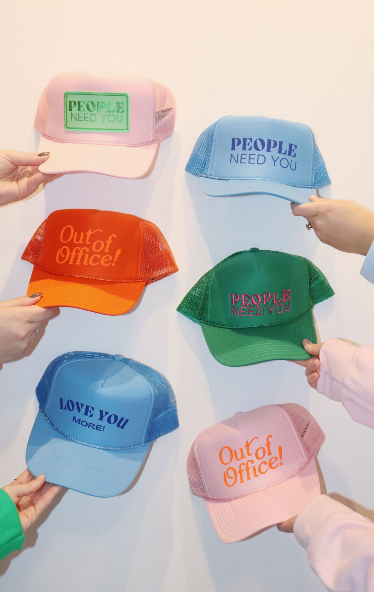 Six colorful caps with various texts held by different hands against a plain background.
