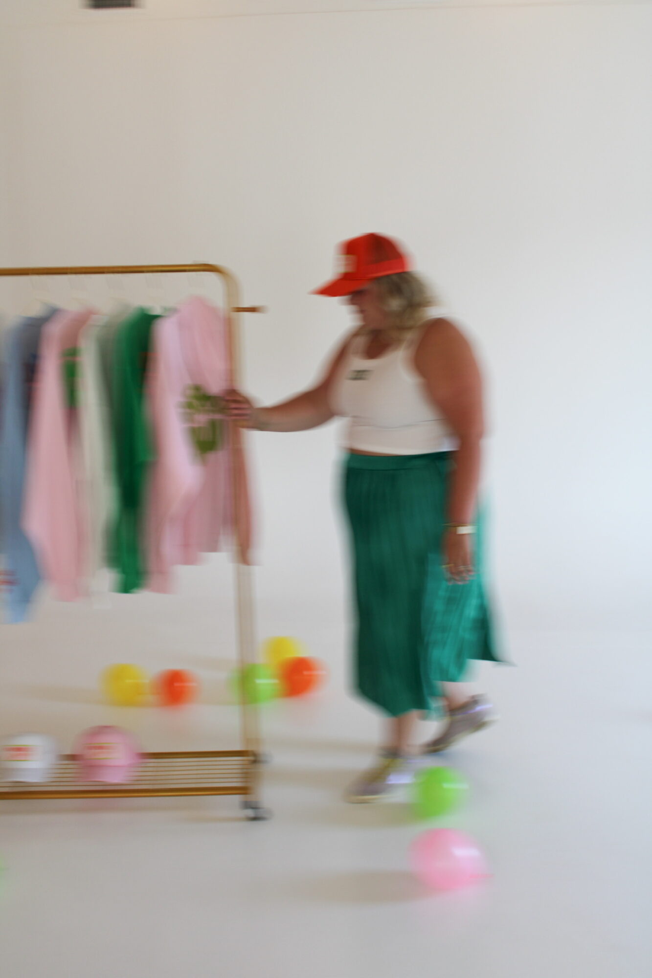 Person wearing a red hat and white top stands next to a clothing rack with colorful clothes, holding a phone.