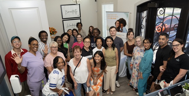 Group of diverse people gathered indoors, smiling for a photo, with a window and framed art in the background.