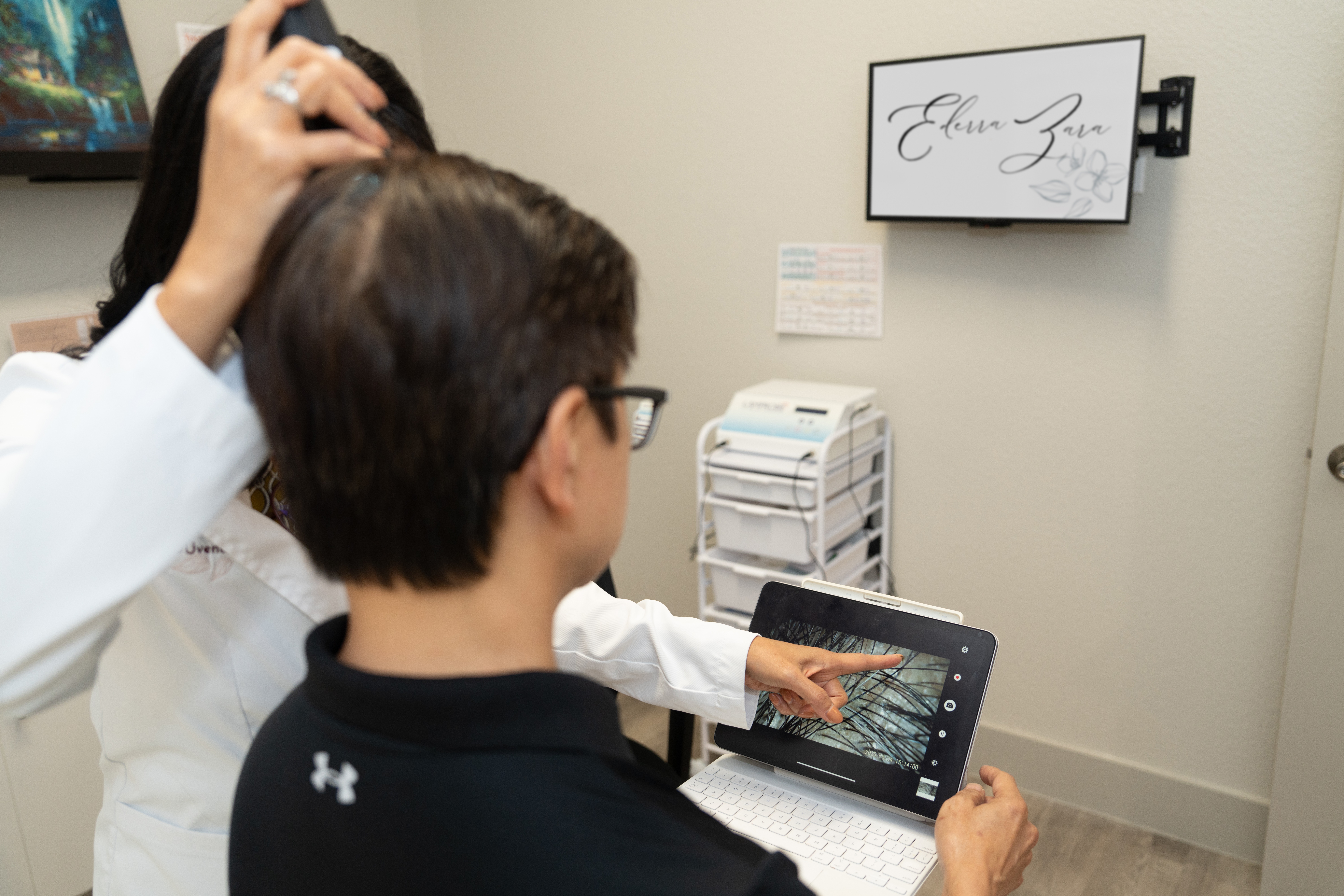 Person with short dark hair and glasses uses a tablet in a medical or office setting, with a monitor on the wall.