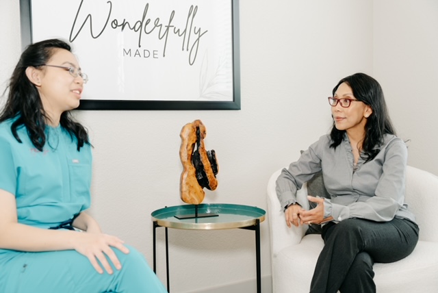 Two women sit in a room with a framed sign that says 'Wonderfully Made'; a small table with a sculpture is between them.