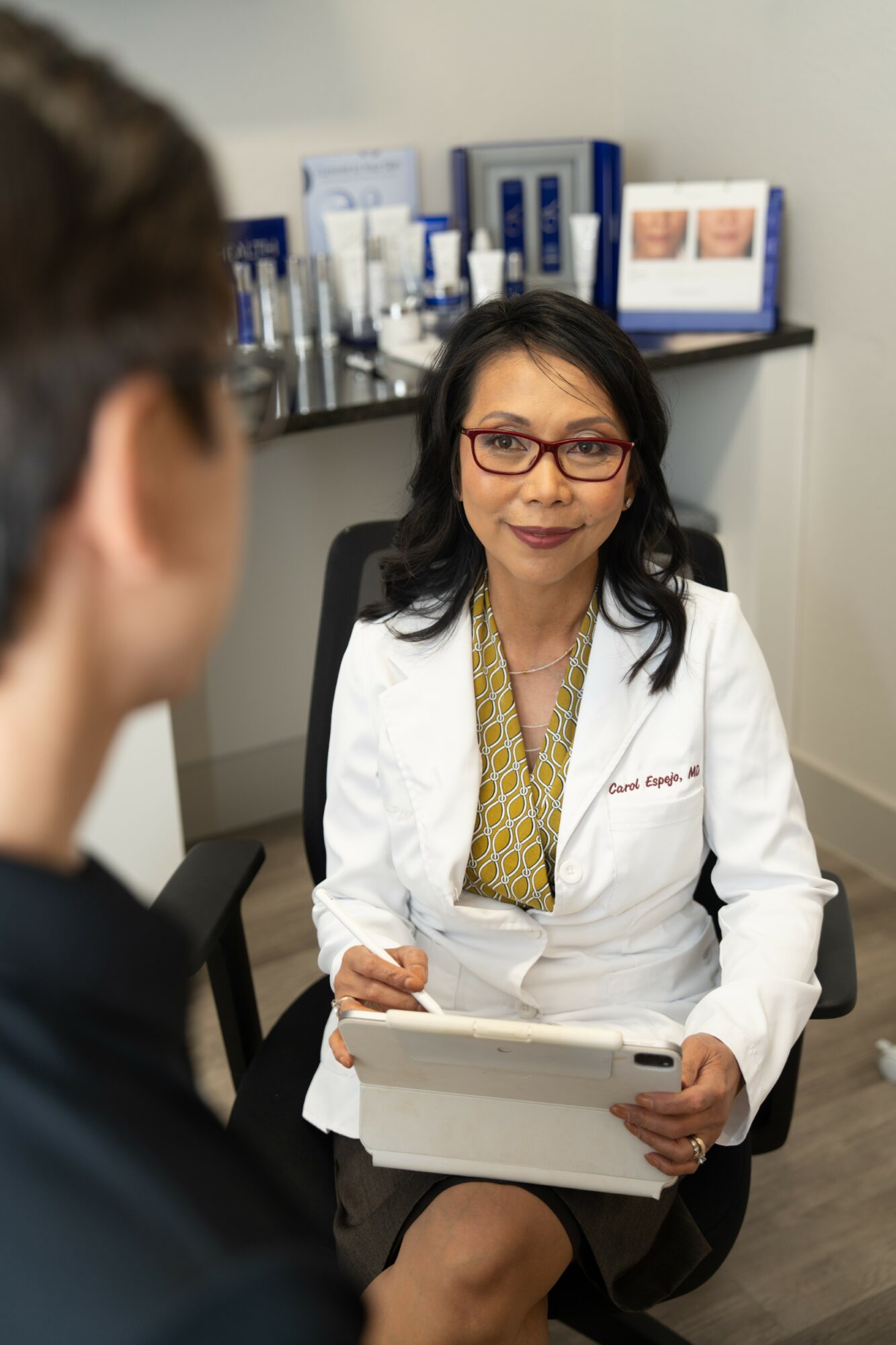 Female doctor with glasses and a white coat sitting in an office, holding a tablet, talking to a patient. Background has medical supplies.