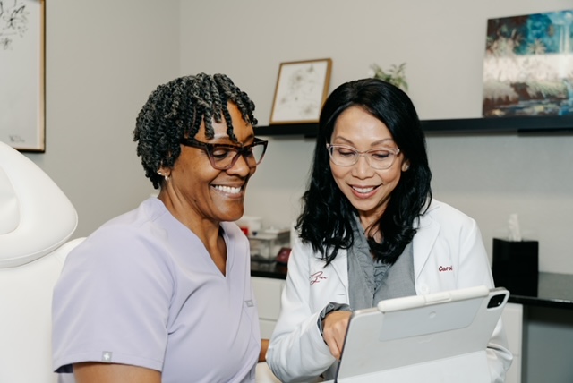 Two women in medical uniforms smiling and looking at a tablet in a clinical setting.