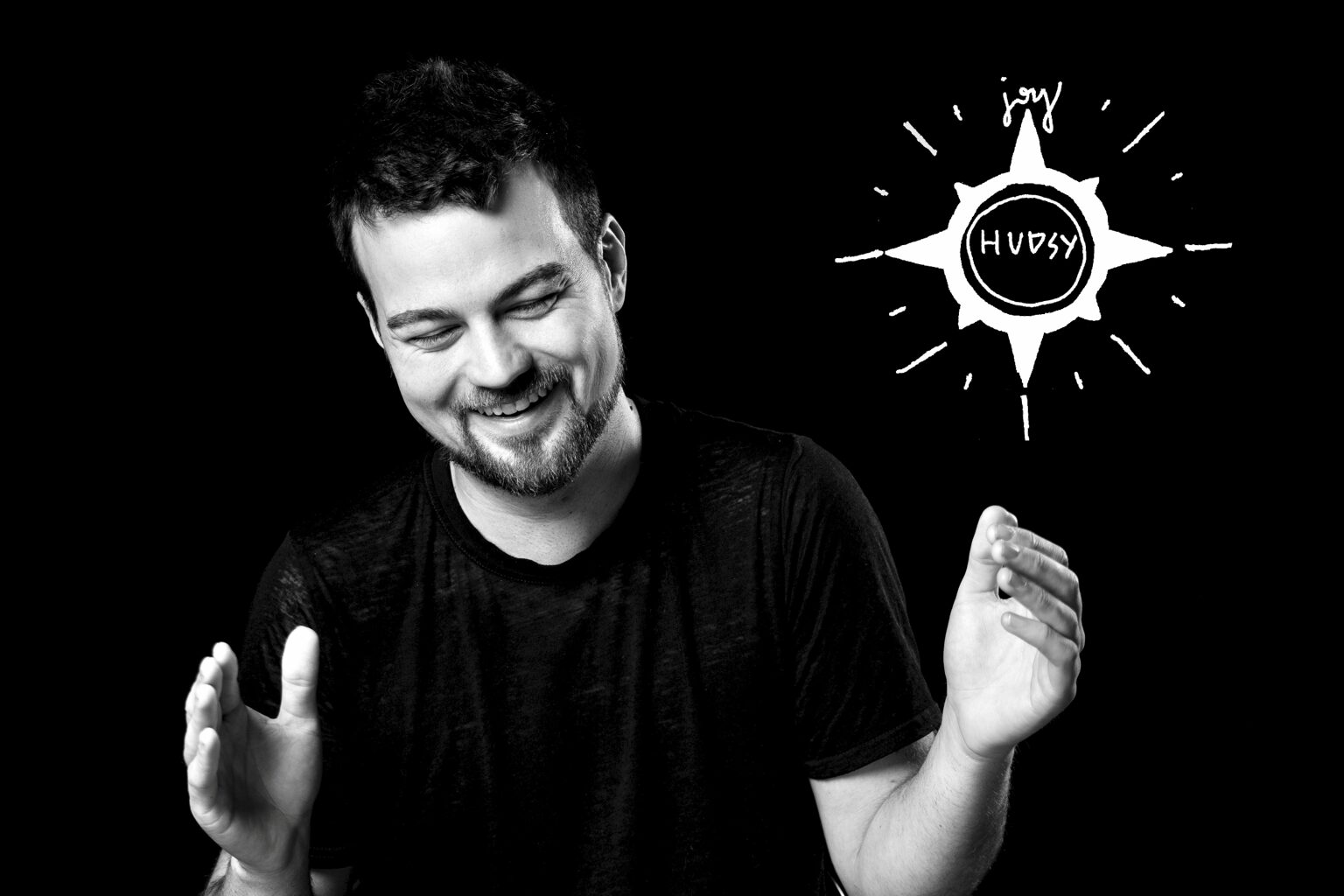 A smiling man with short dark hair and a beard, wearing a black shirt, gestures with his hands against a black background.