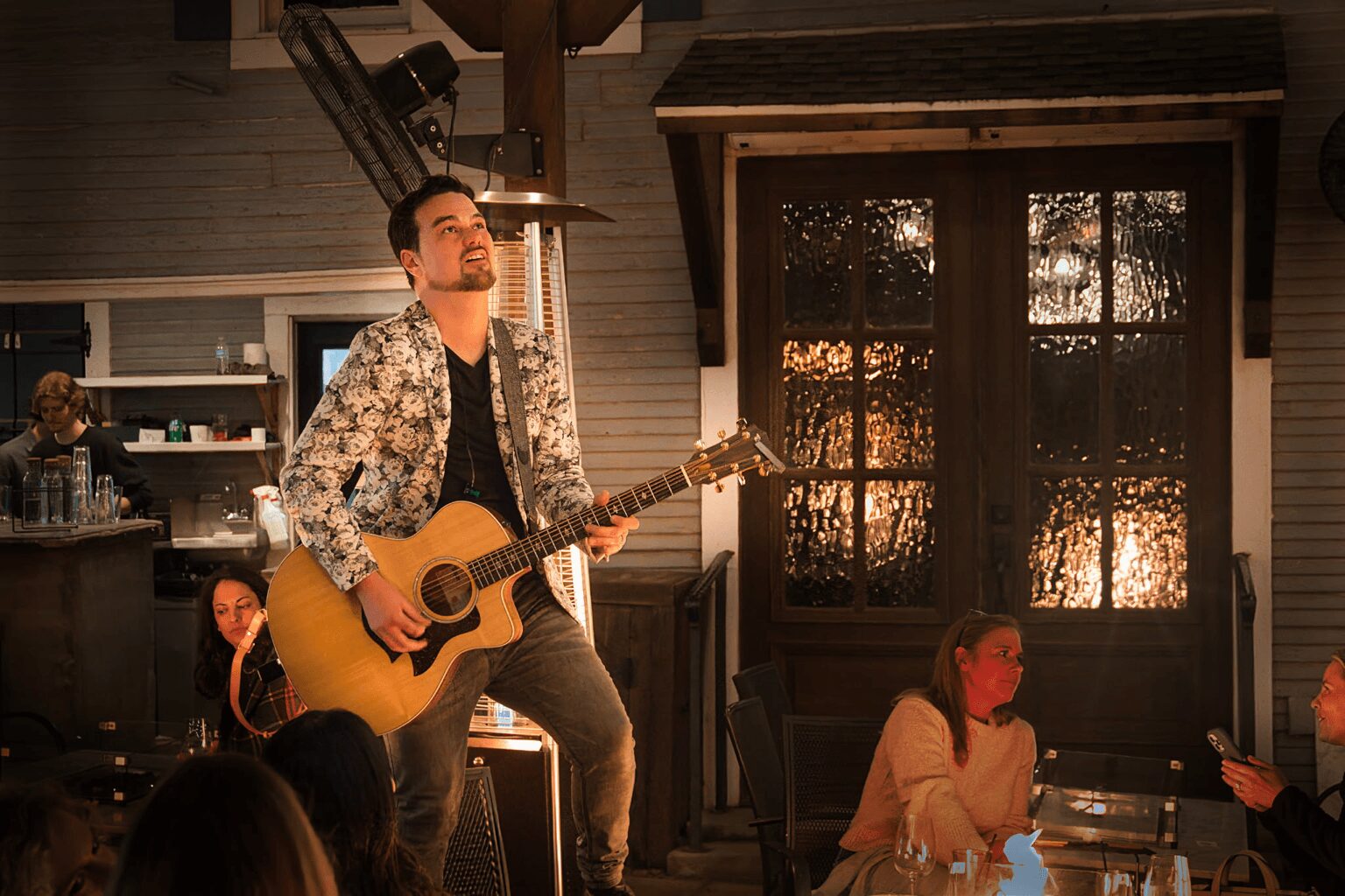 Man playing guitar on stage with audience in front, warm lighting, and a window with curtains in the background.
