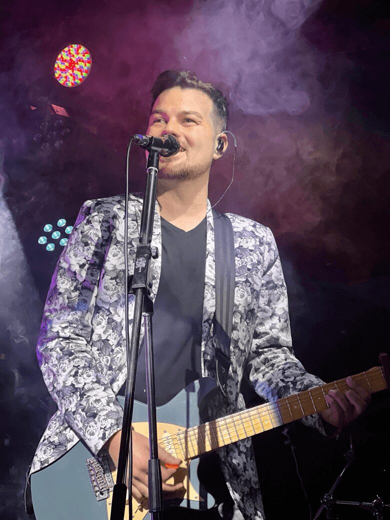 Male musician singing into a microphone and playing an electric guitar on stage with colorful lights and smoke.