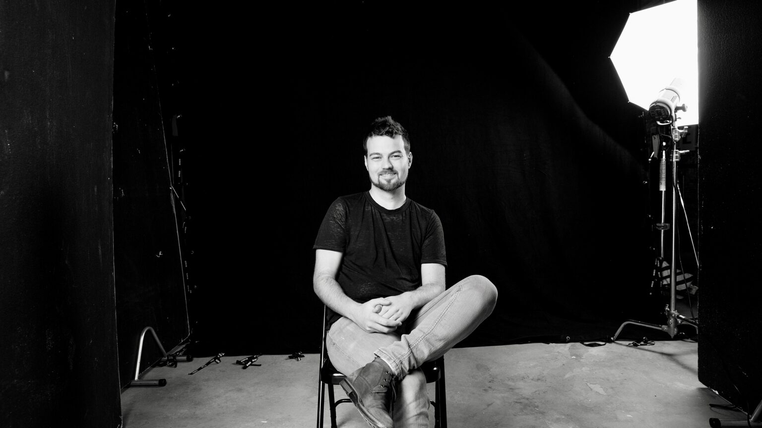 Man sitting on a chair in a studio with a black background and studio light, smiling, wearing a dark t-shirt and jeans.