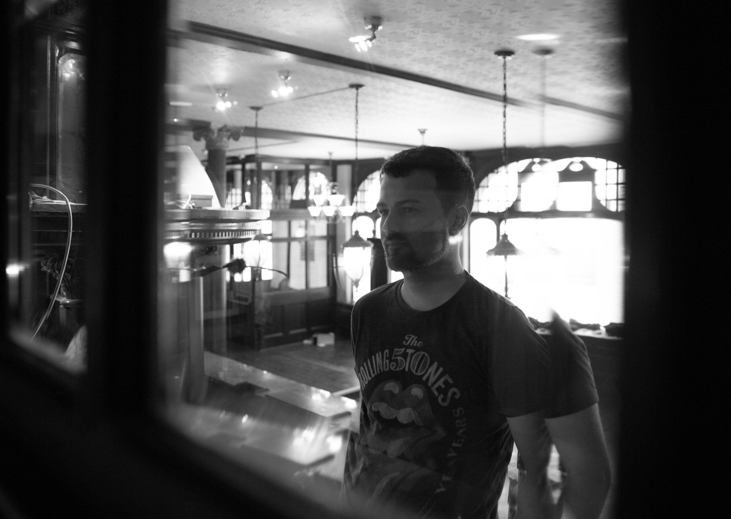 Man with beard and backpack standing inside a cafe or restaurant, seen through a window or glass partition.
